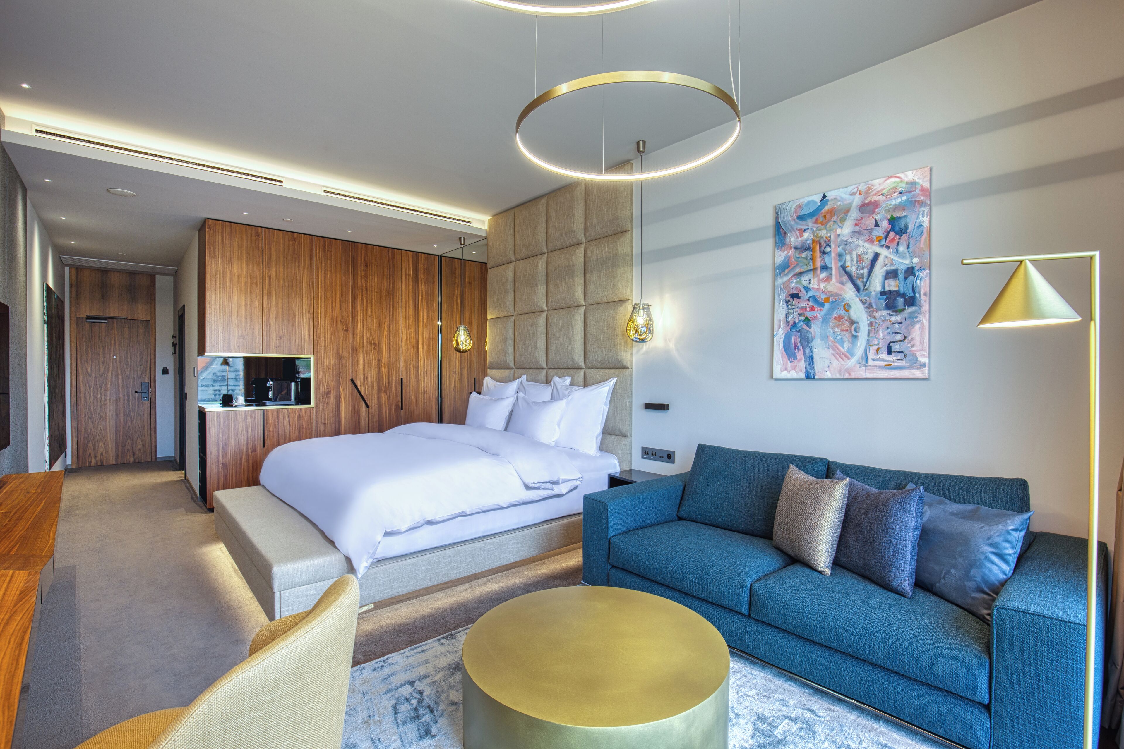 A modern hotel room with a large bed, a sofa, a round coffee table, and a kitchenette.
