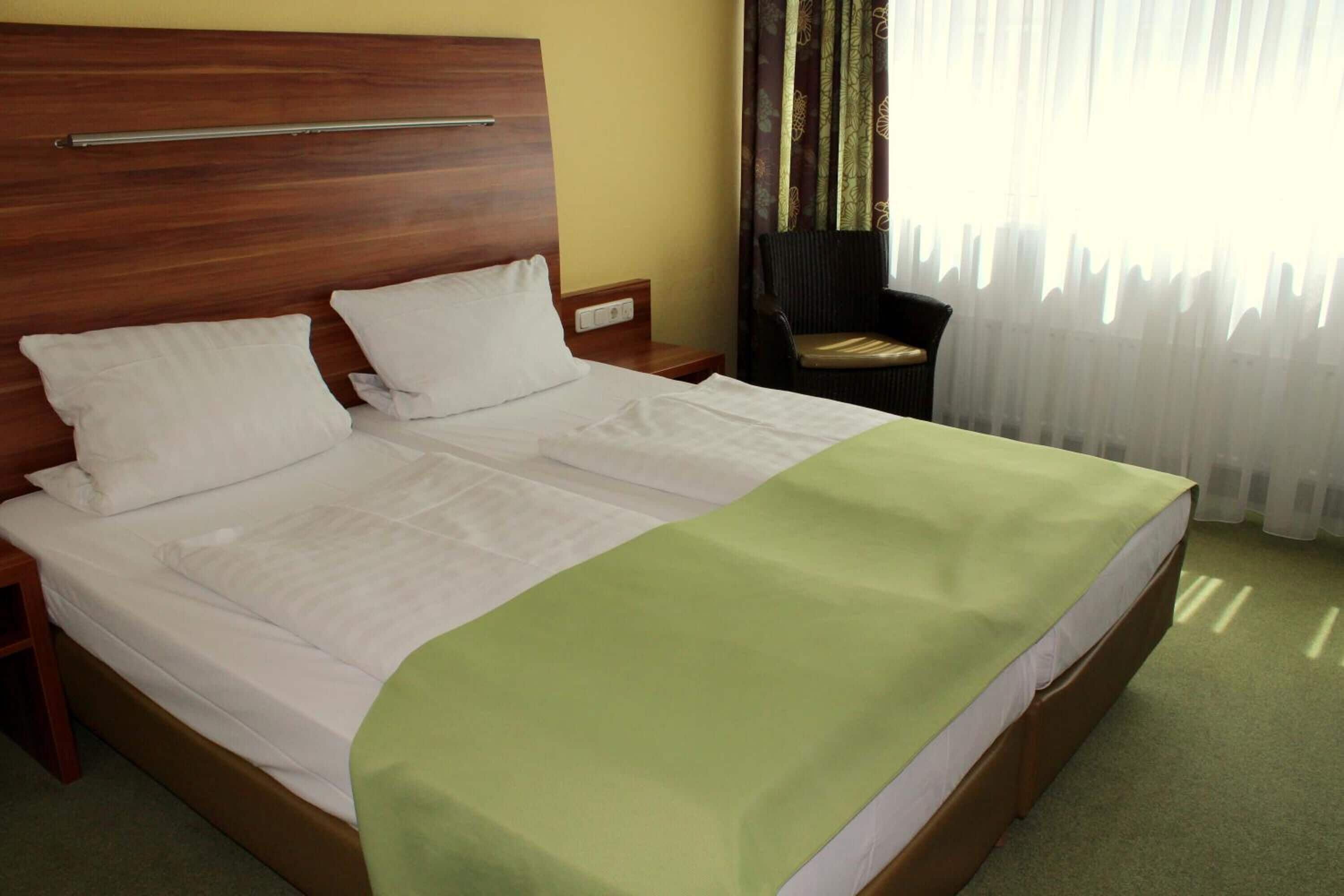 A double bed with a wooden headboard, a green bedspread, a nightstand, a chair, and a window with sheer curtains.