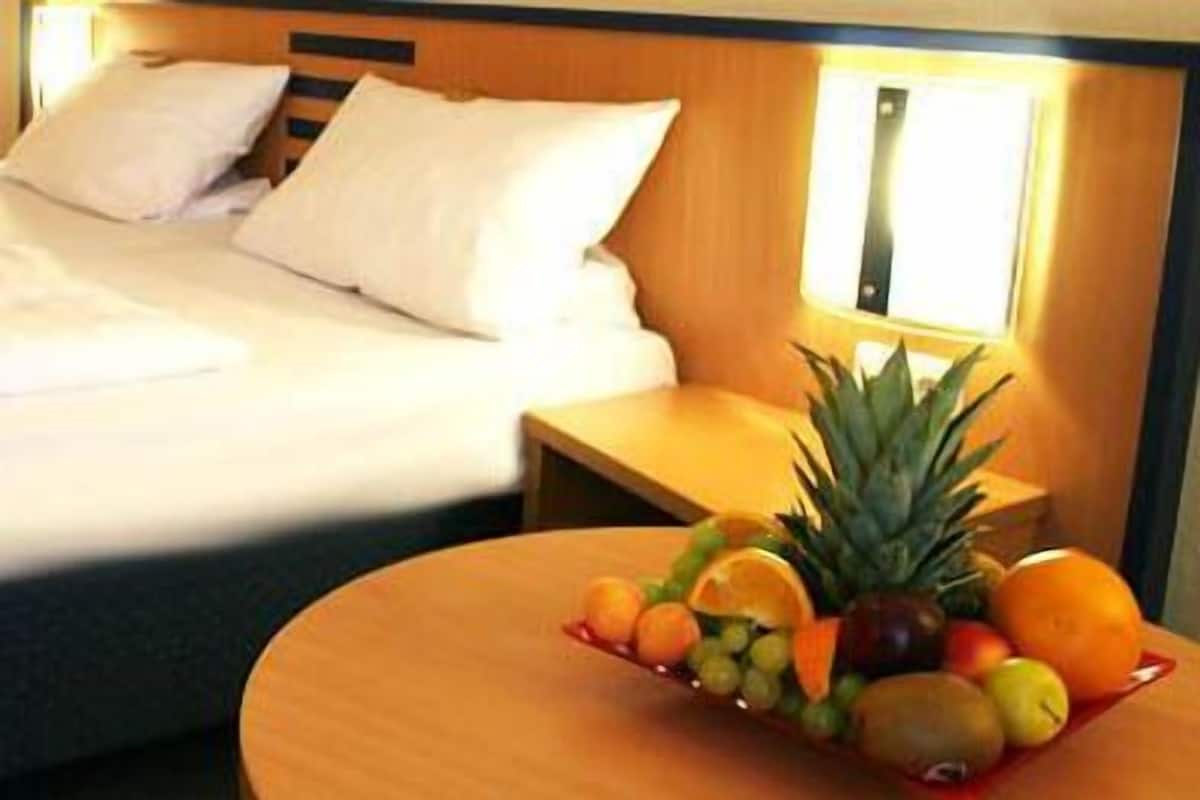 A hotel room with a neatly made bed, a bedside table with a fruit bowl, and a wall-mounted lamp.