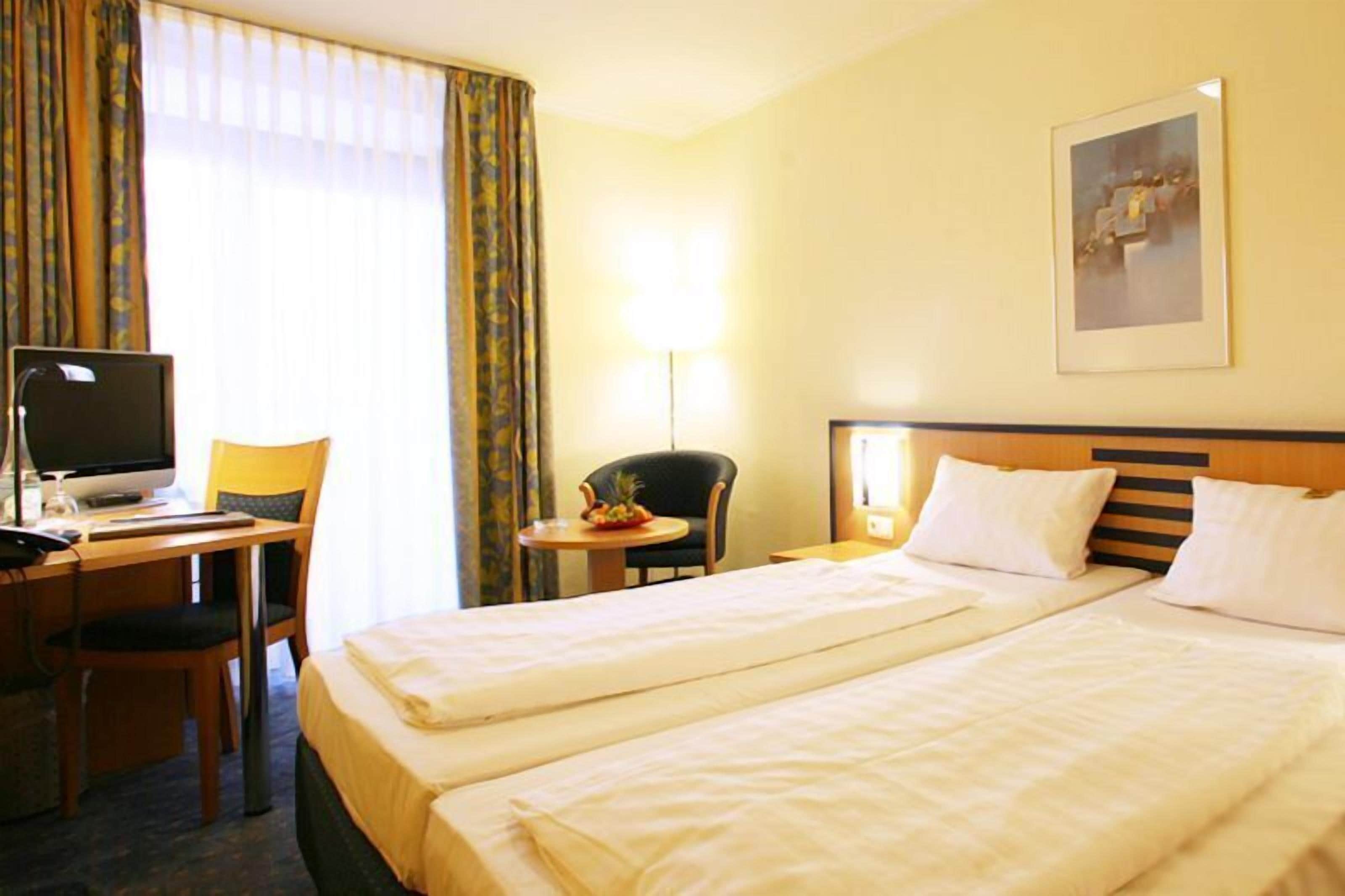 Une chambre d’hôtel avec deux lits, un bureau avec un ordinateur, une chaise, une petite table avec un panier de fruits et une lampe.