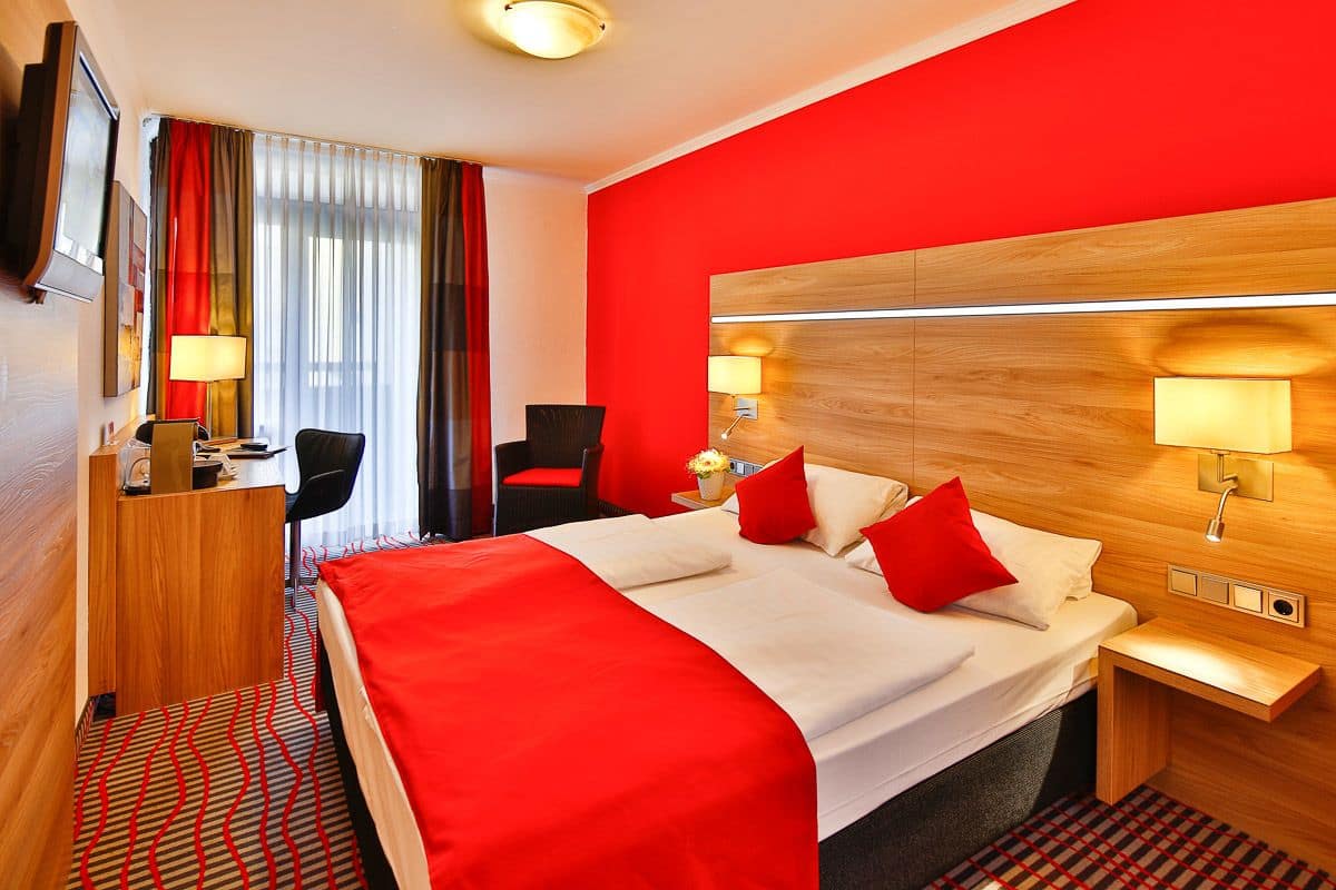 A hotel room with a red accent wall, a large bed with red and white bedding, a desk with a chair, and a TV mounted on the wall.
