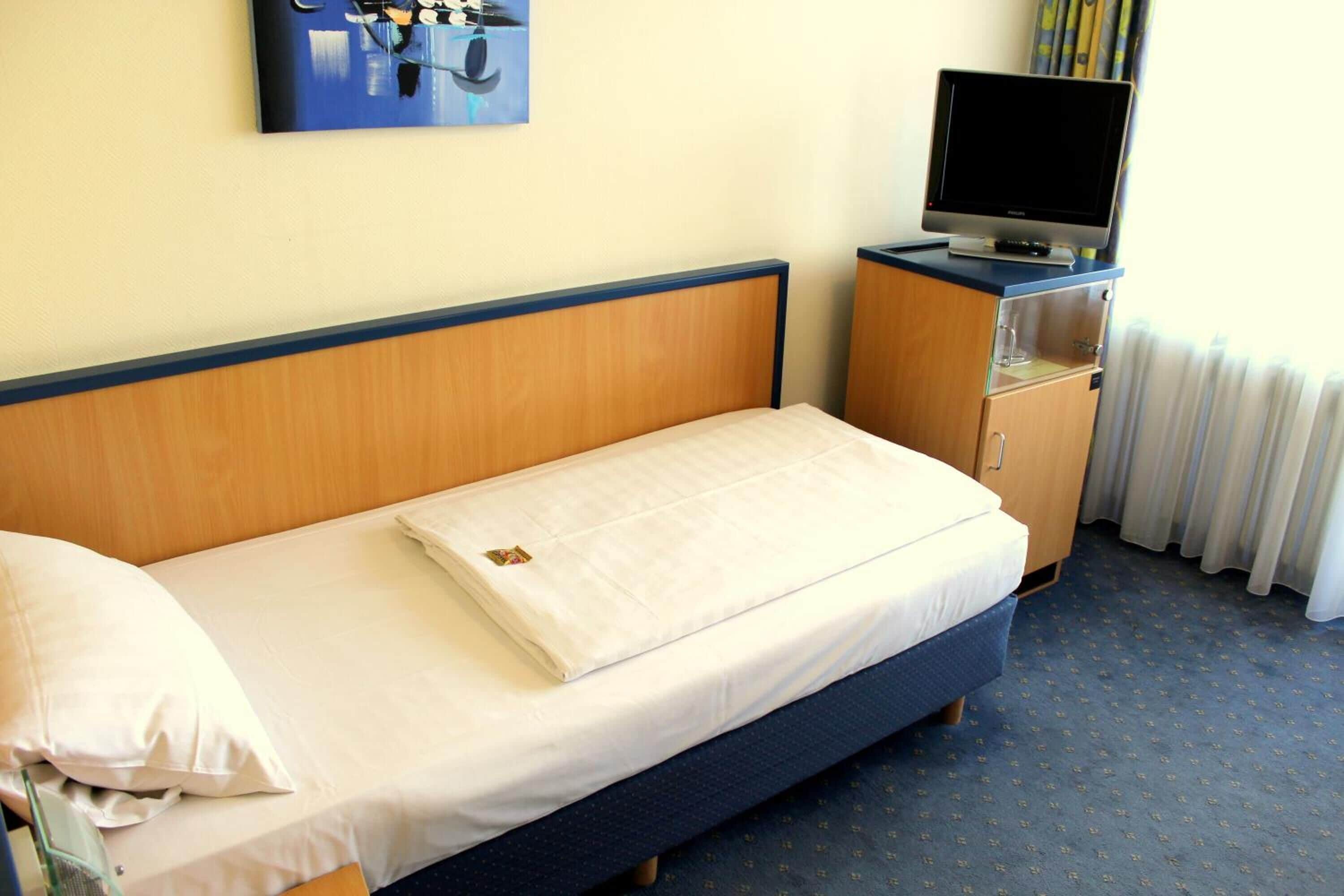 Una cama individual con cabecera de madera, un televisor sobre un mueble bajo y un cuadro en la pared.