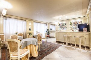 A dining area with a table set for four, surrounded by wicker chairs. A kitchen area with a bar and shelves of items is visible in the background.