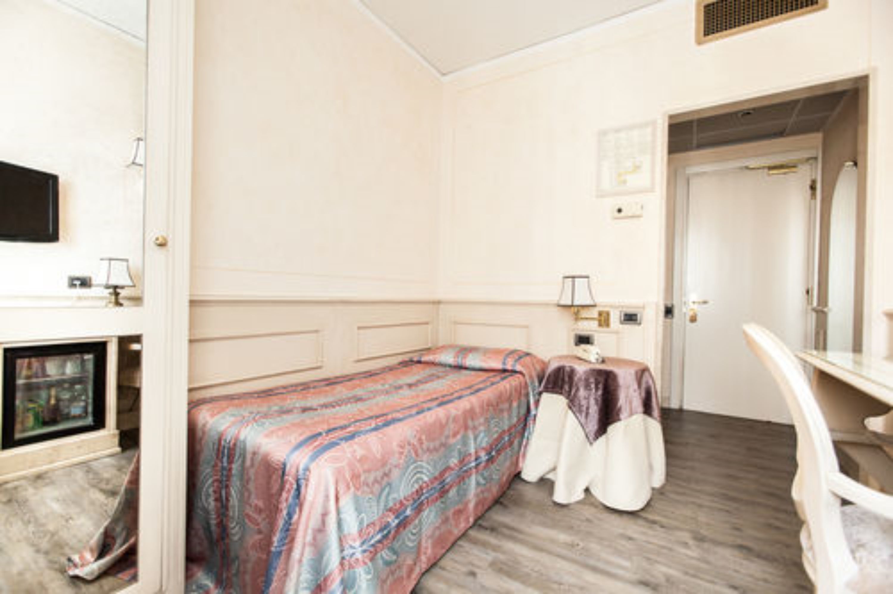 Une pièce comprenant un lit, une petite table avec une lampe, un téléviseur et une porte donnant sur une autre pièce.