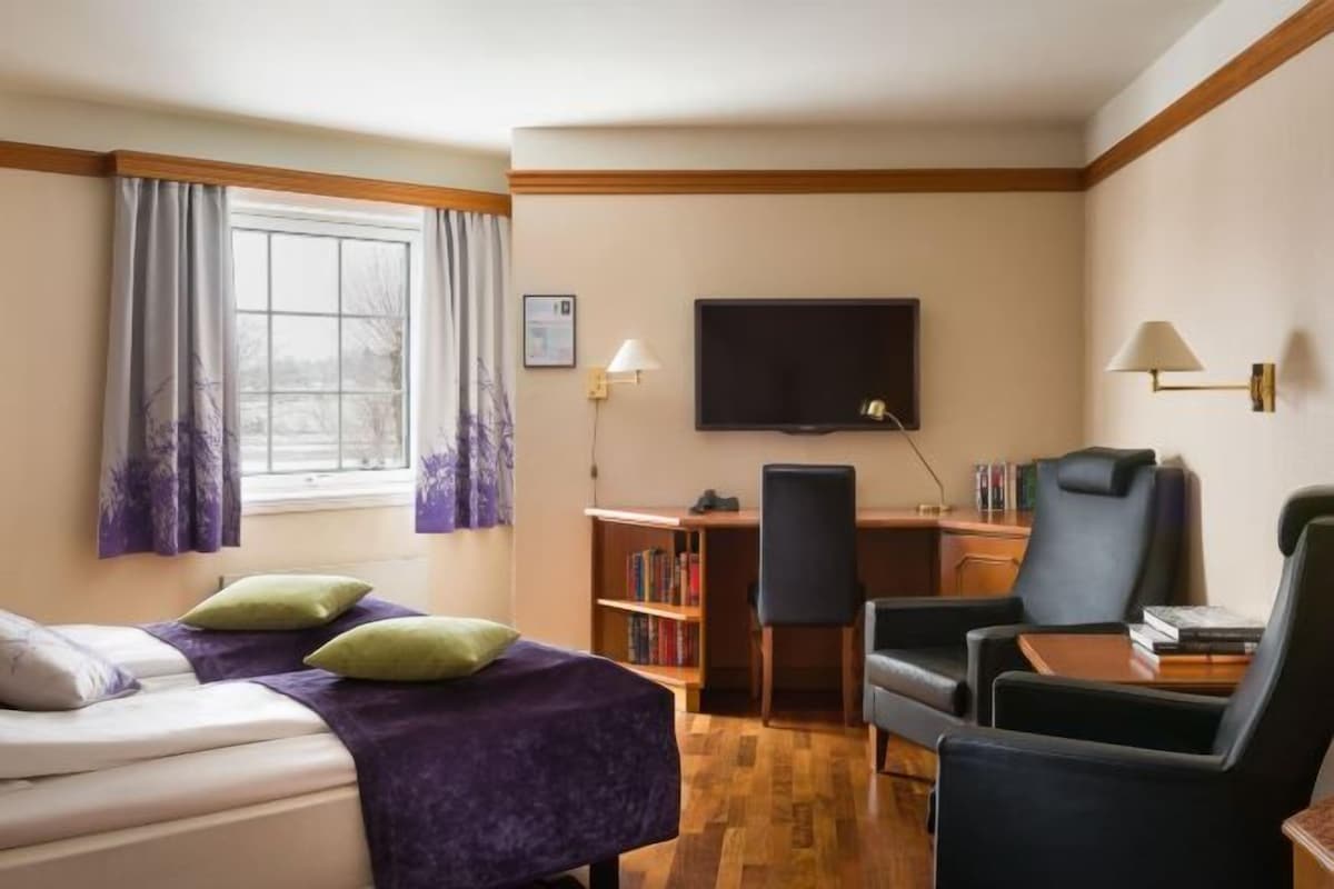 Una habitación de hotel con cama, dos sillas, escritorio, televisor y estantería.
