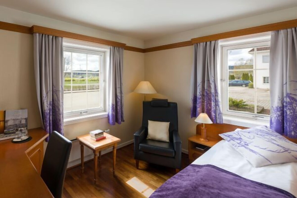 A hotel room with a bed, a desk, a chair, a lamp, and curtains.