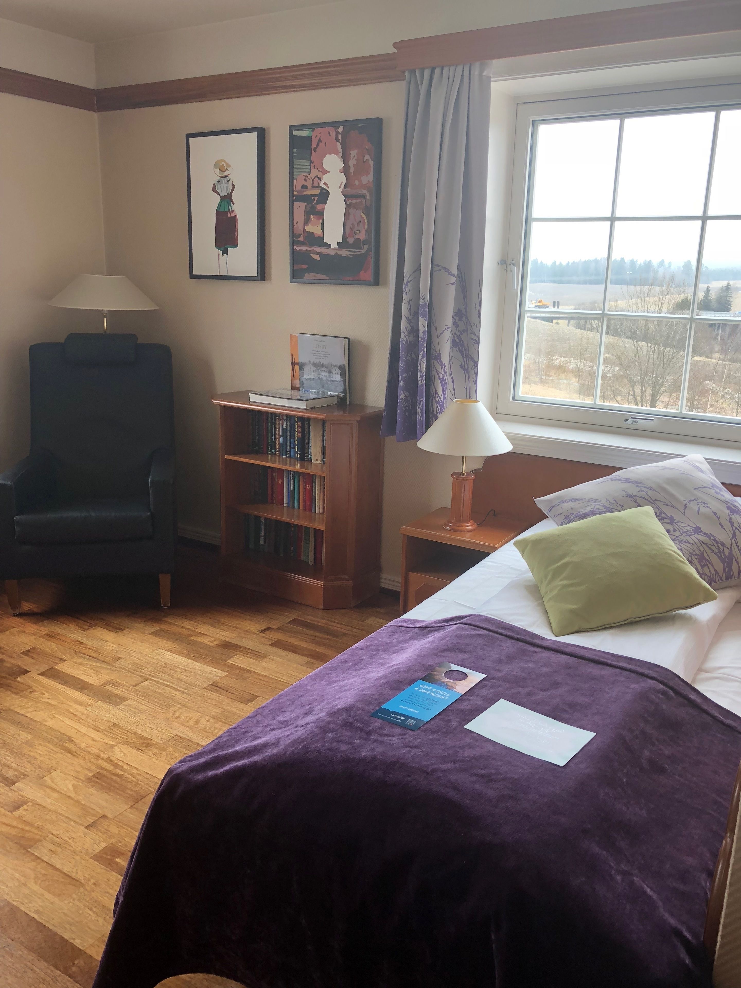 A hotel room with a bed, a chair, a bookshelf, and a window with a view.