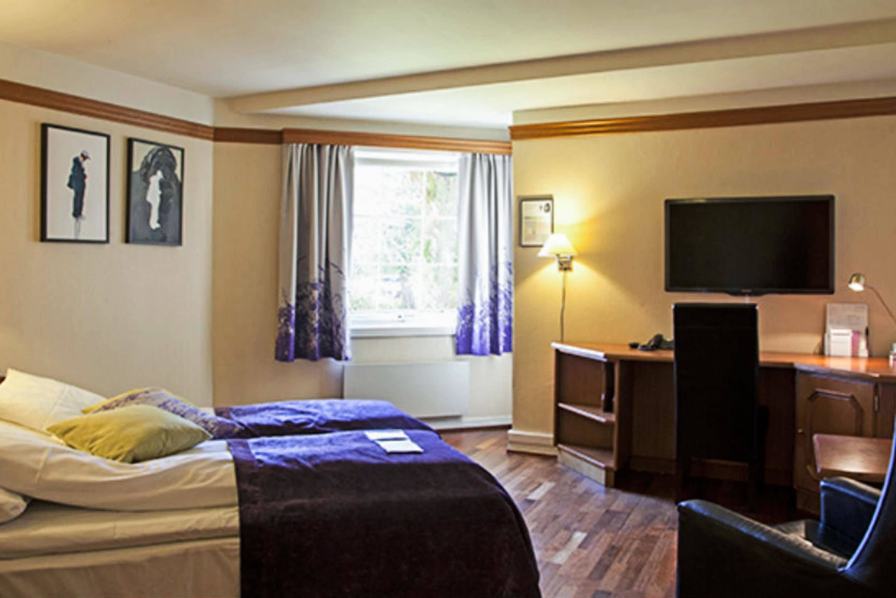 Una habitación de hotel con cama, escritorio, televisión y ventana con cortinas.
