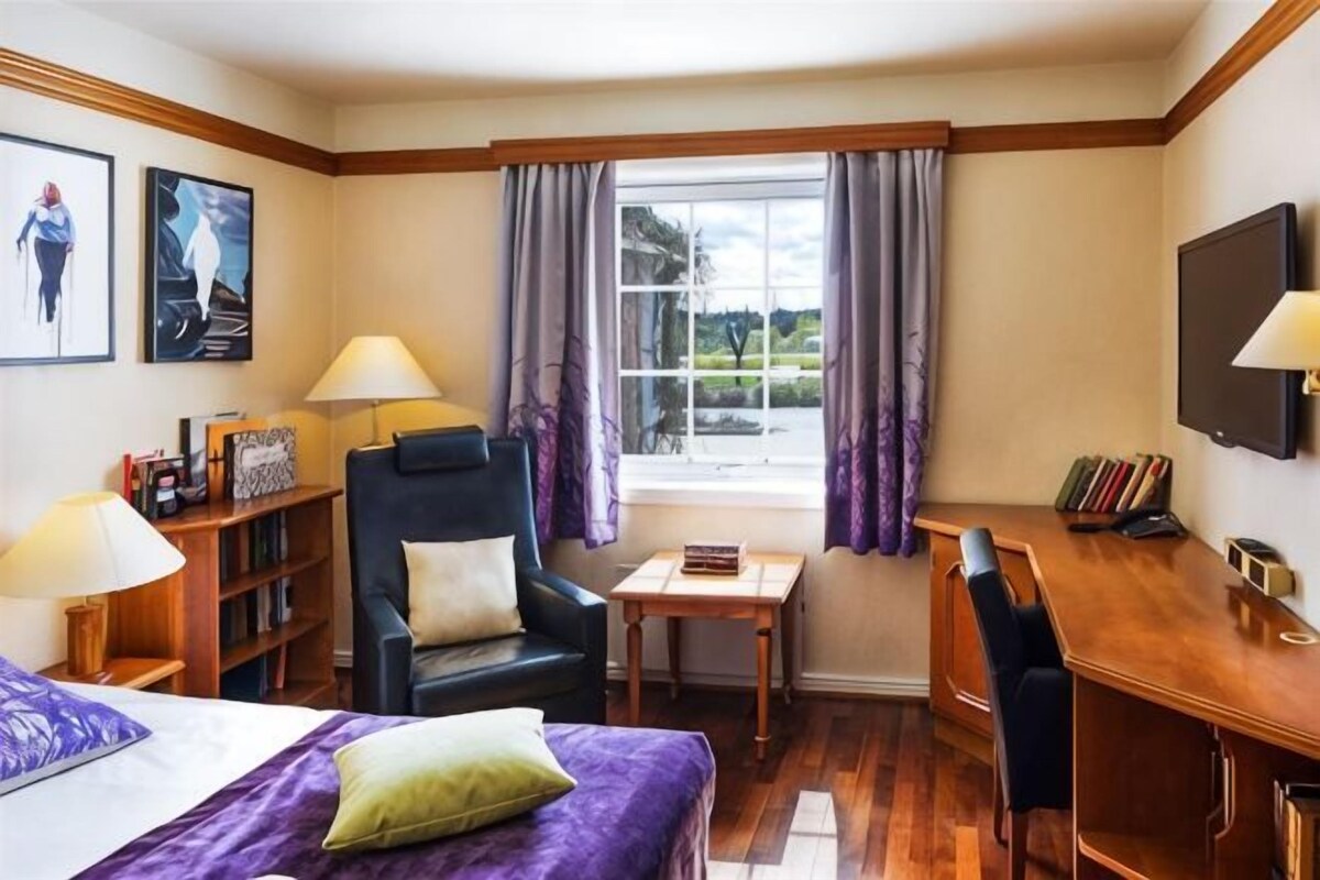 Una habitación de hotel con cama, escritorio, silla, estantería, televisor y ventana con cortinas.