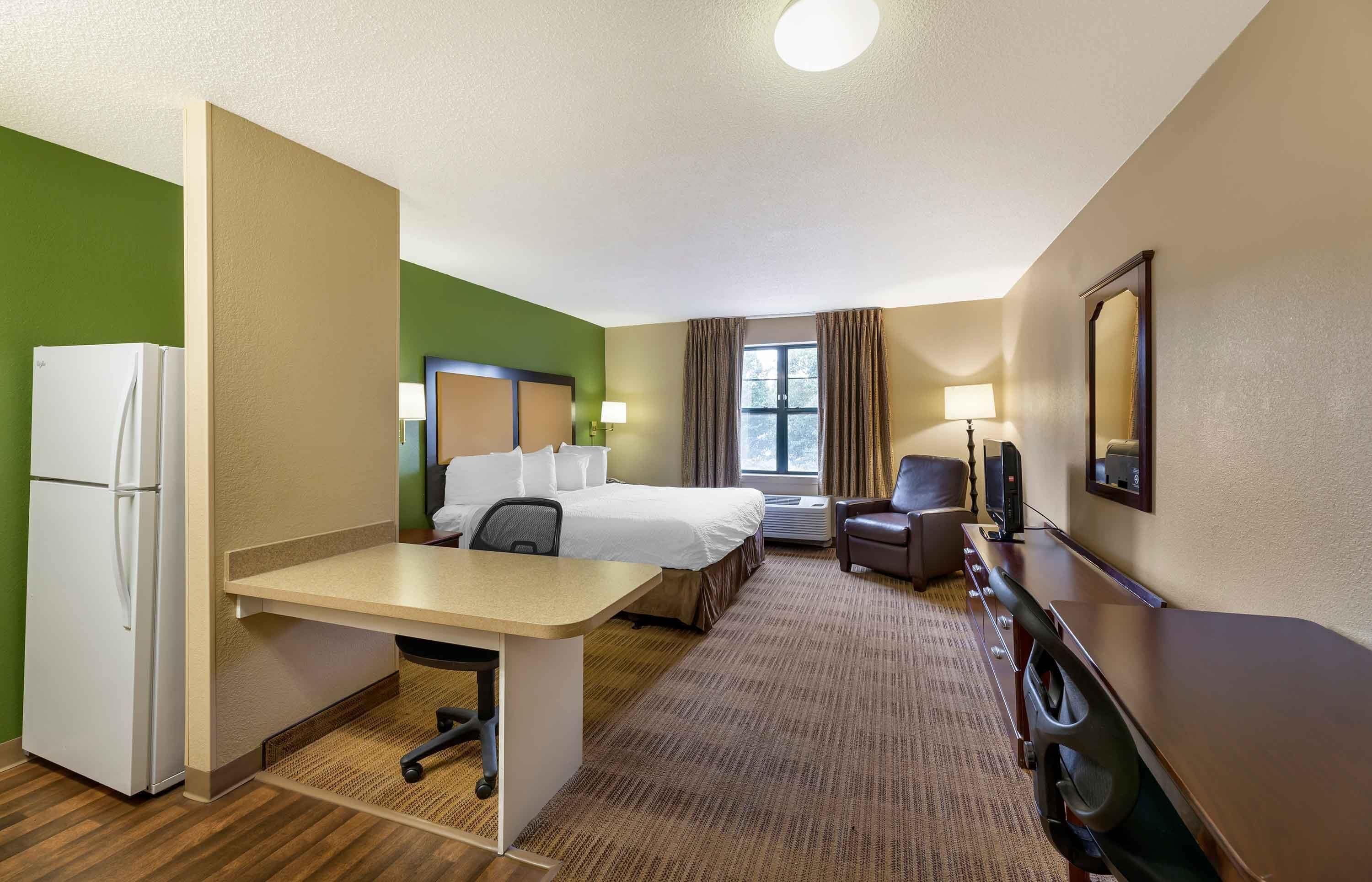 Une chambre d’hôtel équipée d’un lit, d’un bureau, d’une chaise, d’un réfrigérateur et d’une fenêtre avec des rideaux.