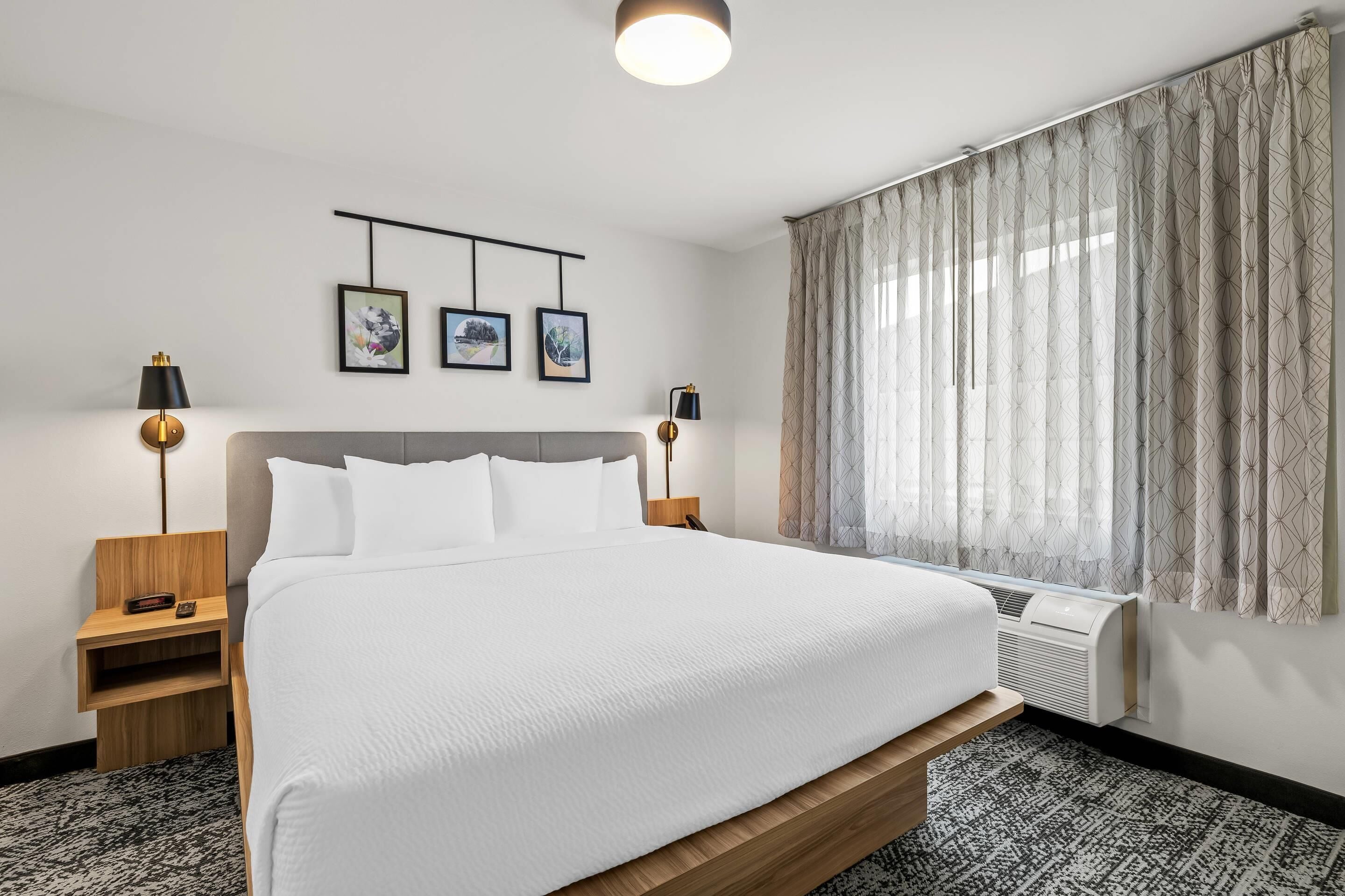 A hotel room with a large bed, two bedside tables, a wall-mounted TV, and a window with curtains.