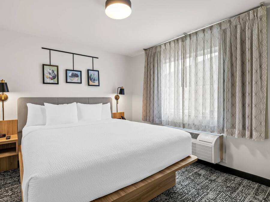 A hotel room with a large bed, two bedside tables, a wall-mounted TV, and a window with curtains.