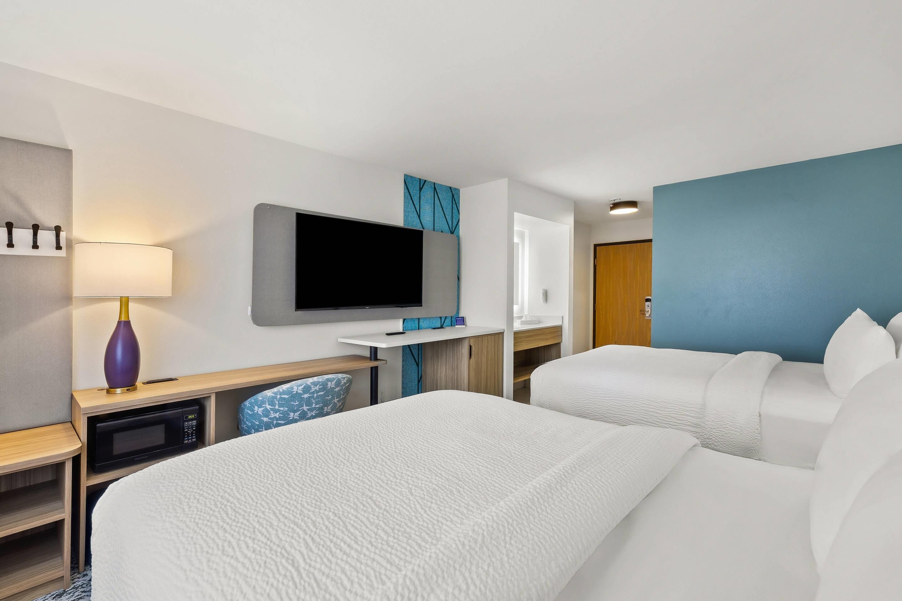 A hotel room with two beds, a desk with a flat-screen TV, a microwave, and a lamp.