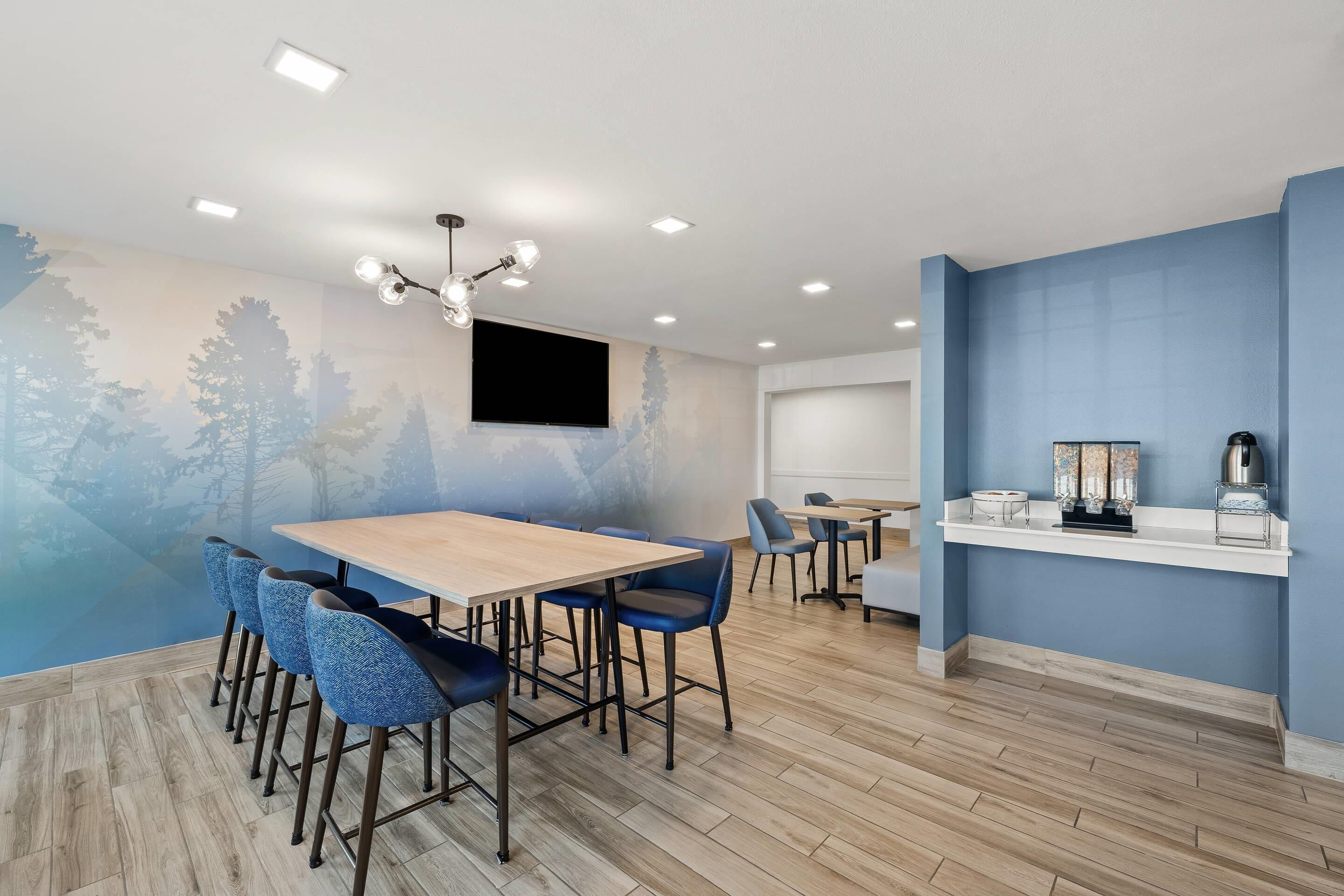 A modern dining area with a wooden table and blue upholstered chairs, a mounted flat-screen TV, and a blue accent wall.