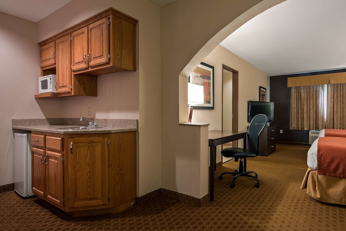 A hotel room with a kitchenette, a desk with a chair, a bed with a red bedspread, and a television.