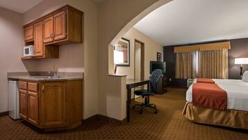 A hotel room with a kitchenette, a desk with a chair, a bed with a red bedspread, and a television.