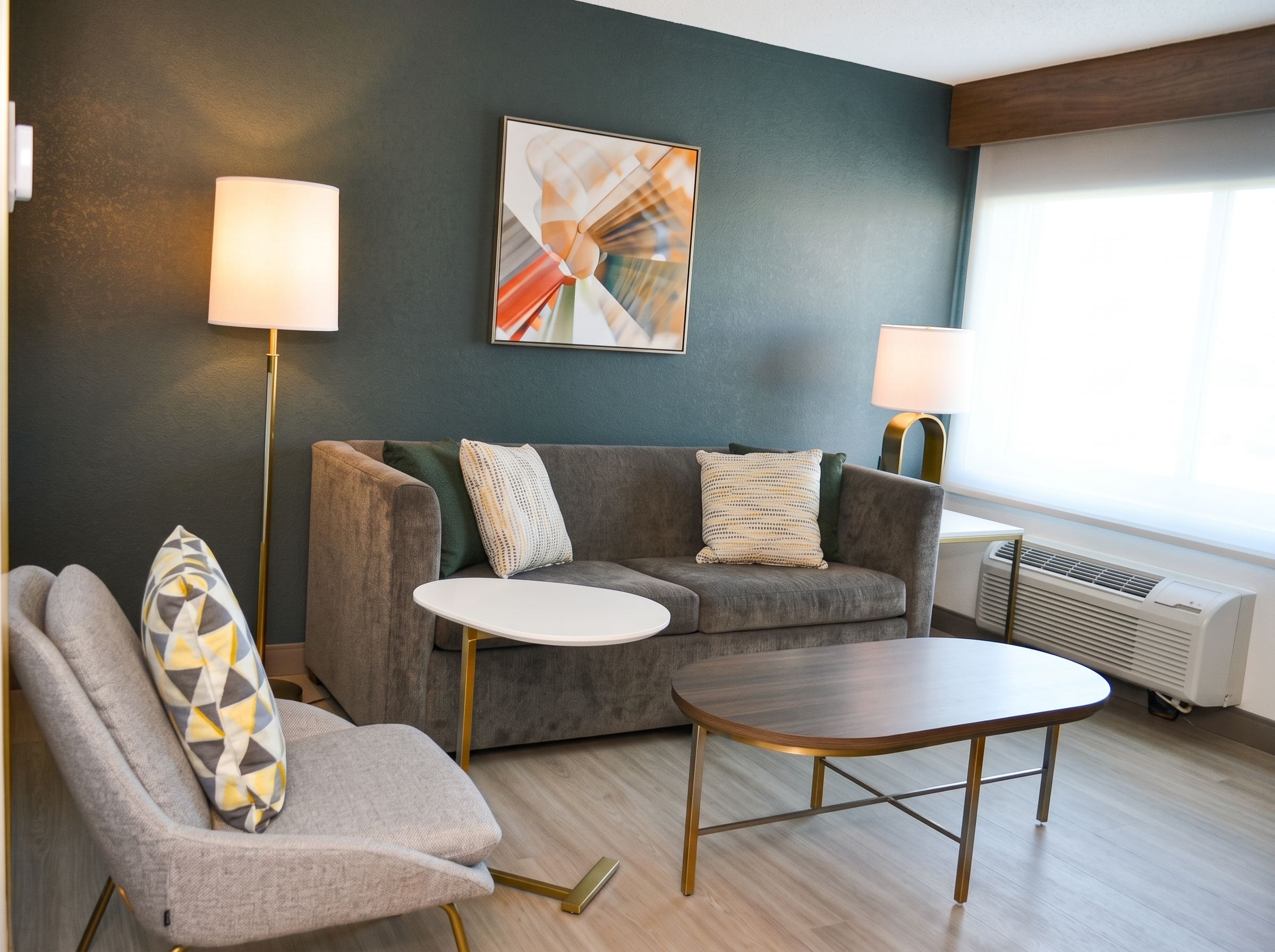 A modern living room with a grey sofa, a round coffee table, and a grey armchair with a patterned cushion. There are two floor lamps and a large abstract painting on the wall.