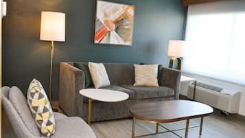 A modern living room with a grey sofa, a round coffee table, and a grey armchair with a patterned cushion. There are two floor lamps and a large abstract painting on the wall.