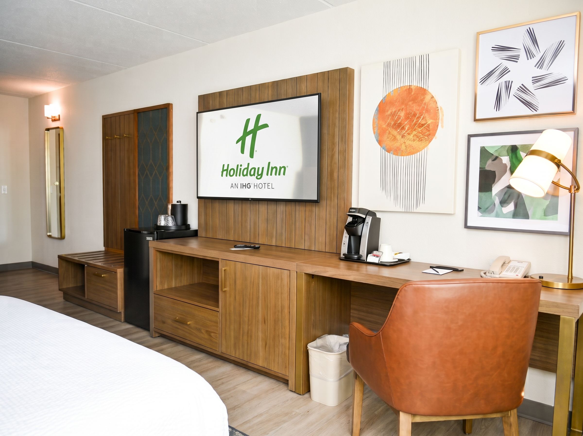 Quarto de hotel com escrivaninha de madeira, uma cama, uma TV exibindo o logotipo da Holiday Inn, uma máquina de café e obras de arte na parede.