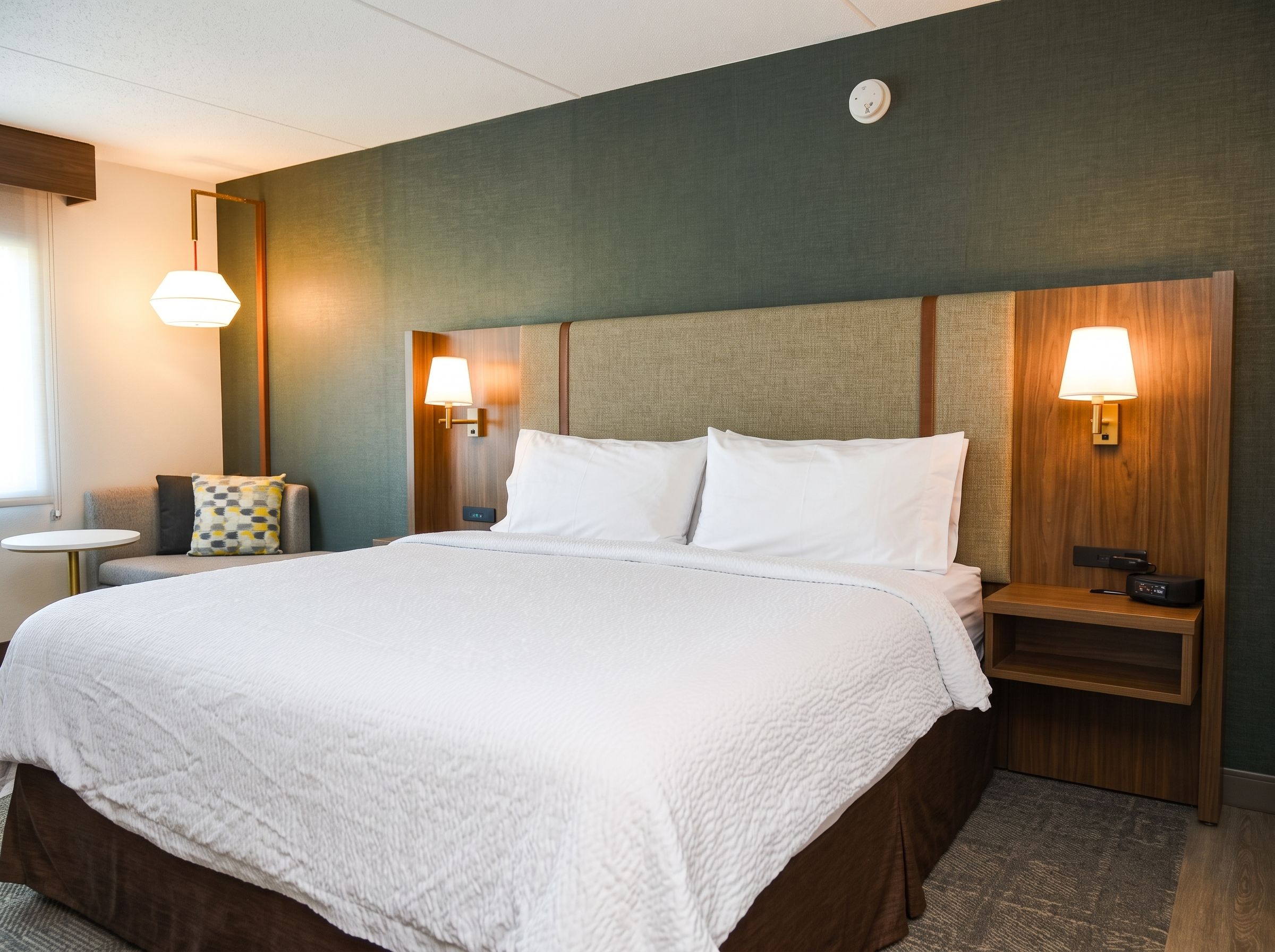 Une chambre d’hôtel avec un grand lit, deux tables de chevet avec des lampes, une petite table et un luminaire fixé au mur.