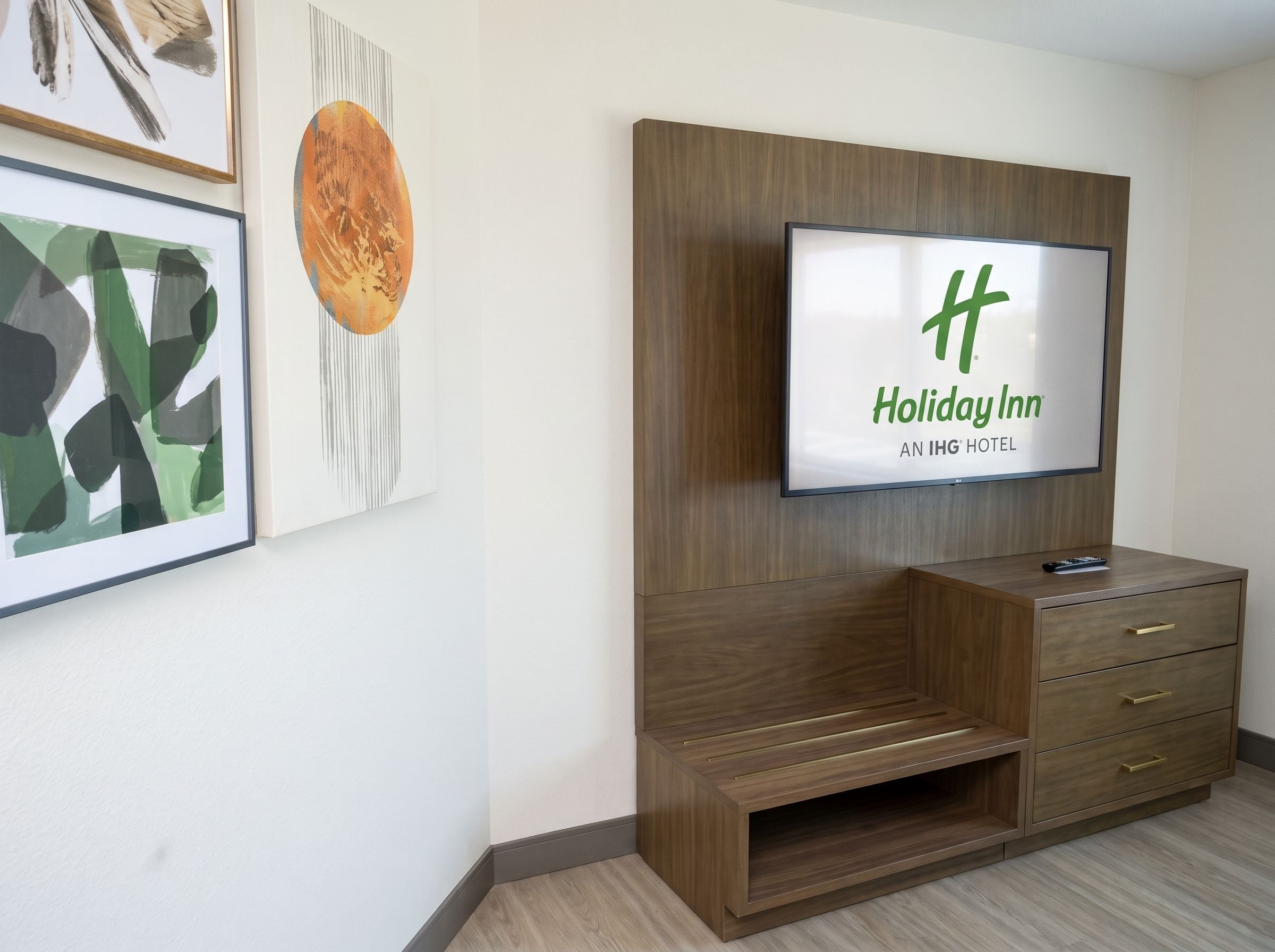 A Holiday Inn hotel room with a flat-screen TV mounted on a wooden cabinet, a small TV stand, and a framed artwork on the wall.