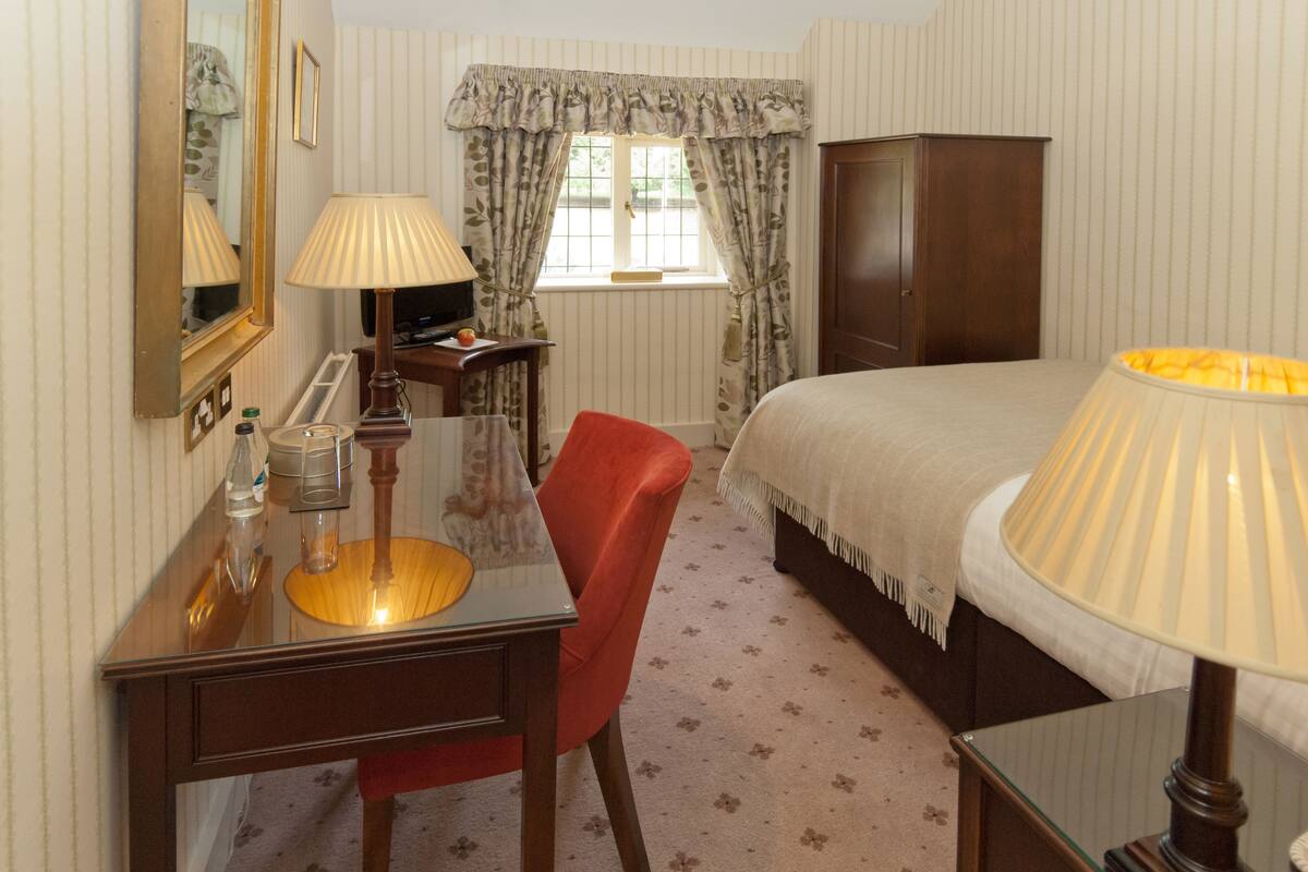 Ein Schlafzimmer mit einem Bett, einem Schreibtisch mit Lampe, einem Stuhl, einem Kleiderschrank und einem Fenster mit Vorhängen.