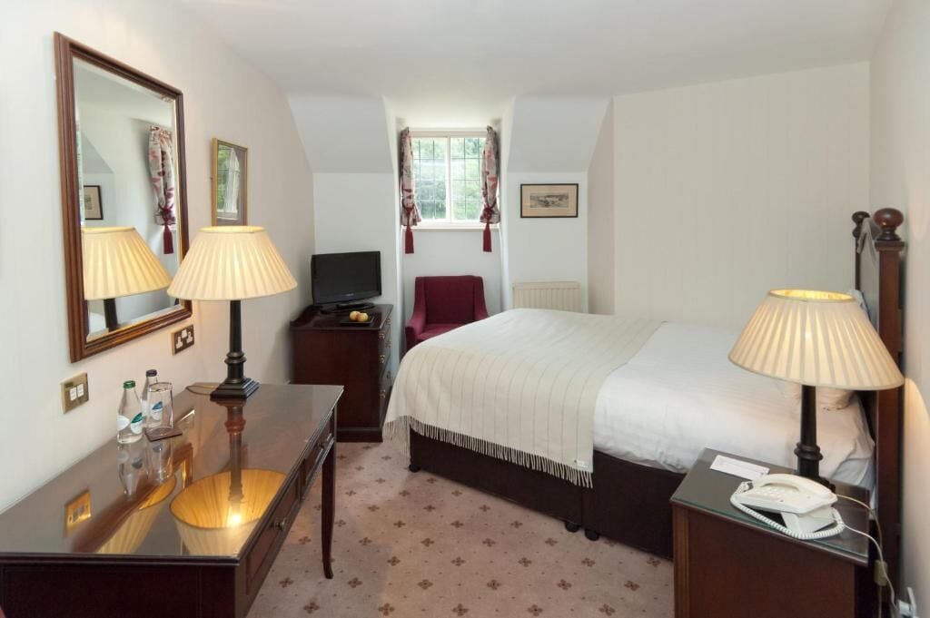 A hotel room with a bed, bedside lamps, a desk with a lamp, a chair, a TV, a telephone, and a window with curtains.
