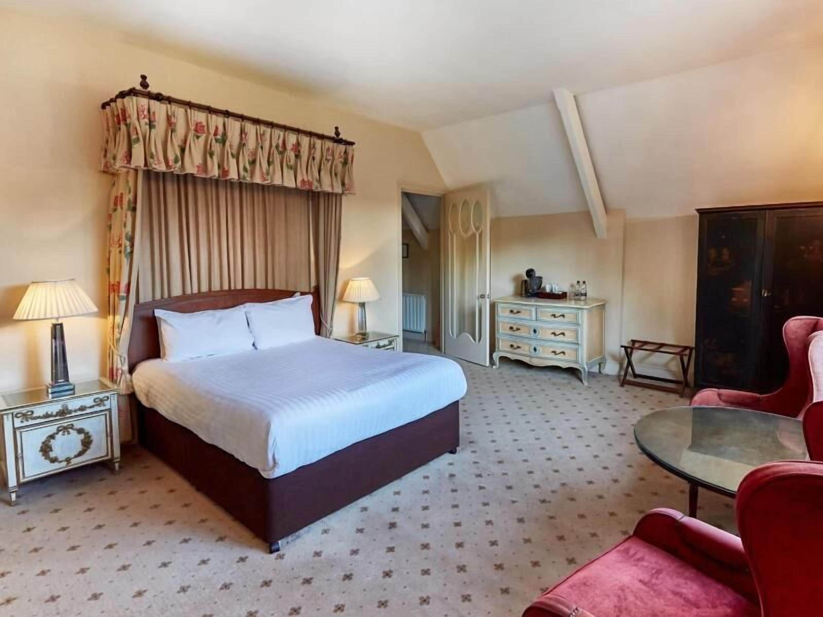 A hotel room with a large bed, two bedside tables, a dresser, a glass table, and a red chair.