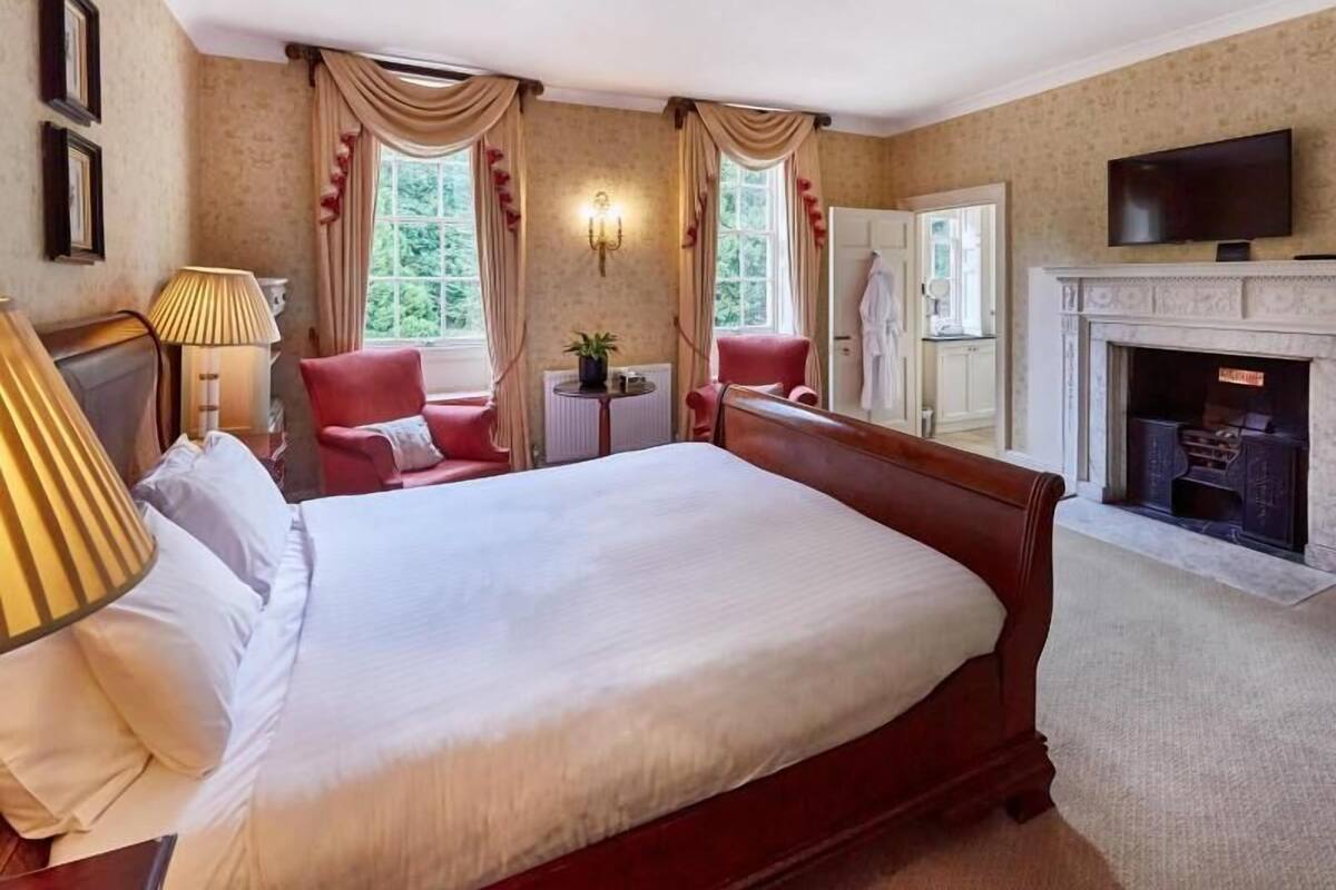 Ein Hotelzimmer mit einem großen Bett, zwei Sesseln, einem kleinen Tisch mit Lampe, einem Kamin und einem Fernseher.