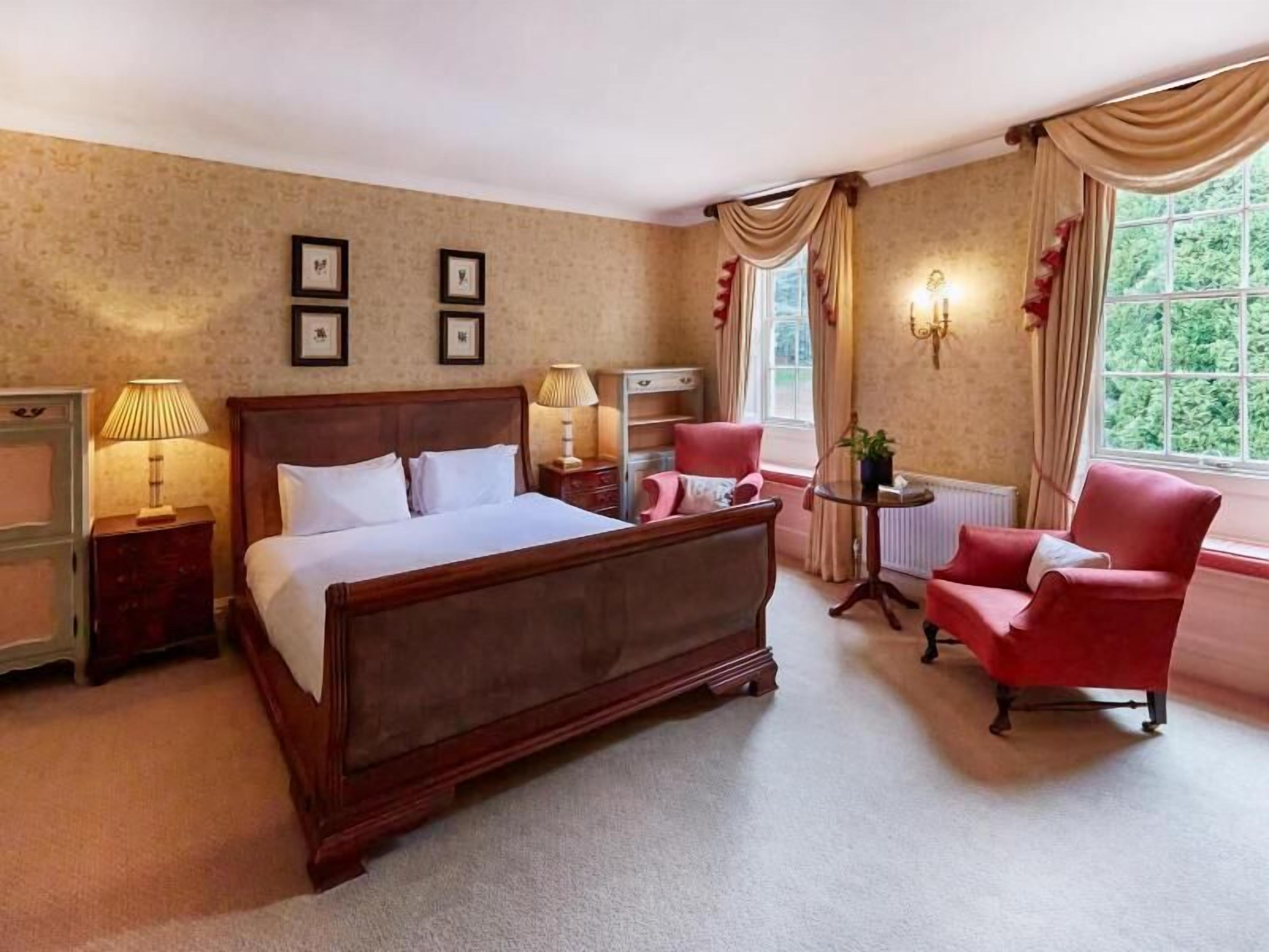 Habitación de hotel con una cama grande, dos mesitas de noche con lámparas, una silla, una pequeña mesa con una planta y una ventana con cortinas.