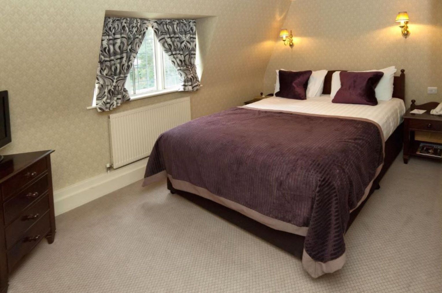 A bedroom with a large bed, two bedside lamps, a dresser, and a window with patterned curtains.