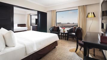 Habitación de hotel con una cama grande, un escritorio con silla, televisión y vistas al aeropuerto.