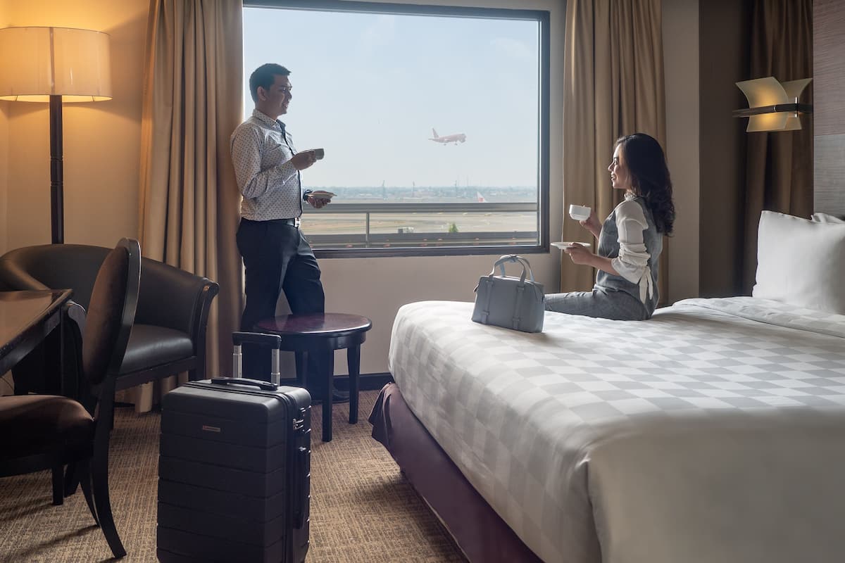 Una habitación de hotel con un ventanal grande, una cama, un escritorio, una silla y una maleta.