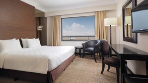A hotel room with a large bed, a desk, a chair, a television, and a window with a view of the airport. - Jakarta Airport Hotel (Tangerang)
