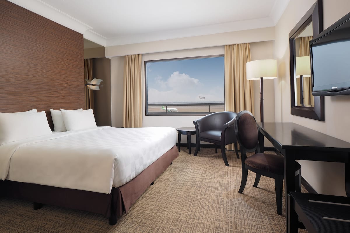 Habitación de hotel con una cama grande, un escritorio, una silla, un televisor y una ventana con vistas al aeropuerto.