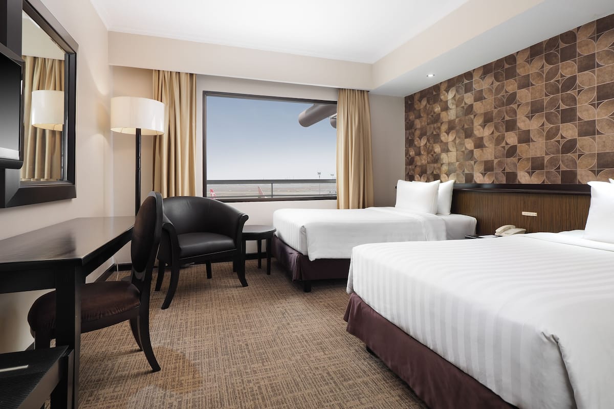 Habitación de hotel con dos camas, un escritorio, una silla y un amplio ventanal con vistas al aeropuerto.