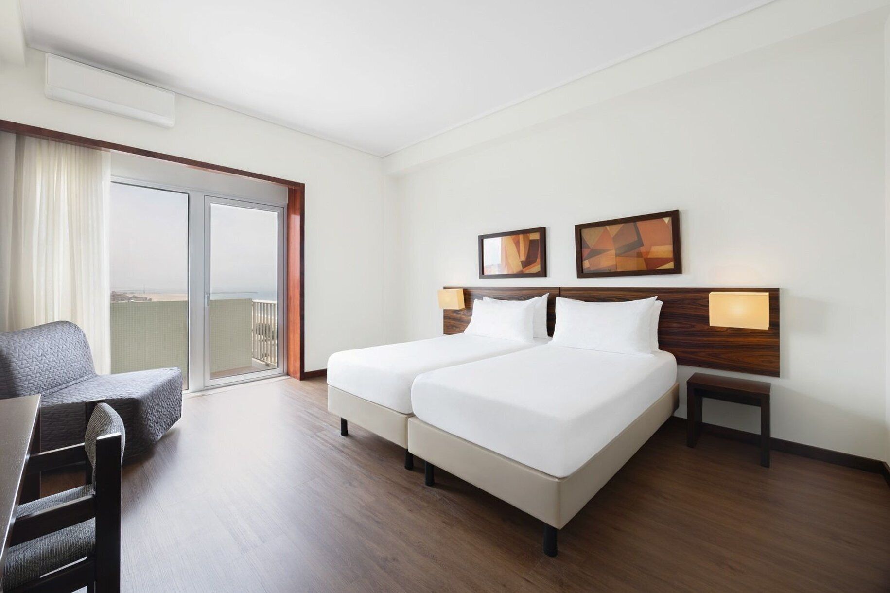 Habitación de hotel con dos camas, un escritorio, una silla, un balcón con vistas y dos lámparas de pared.
