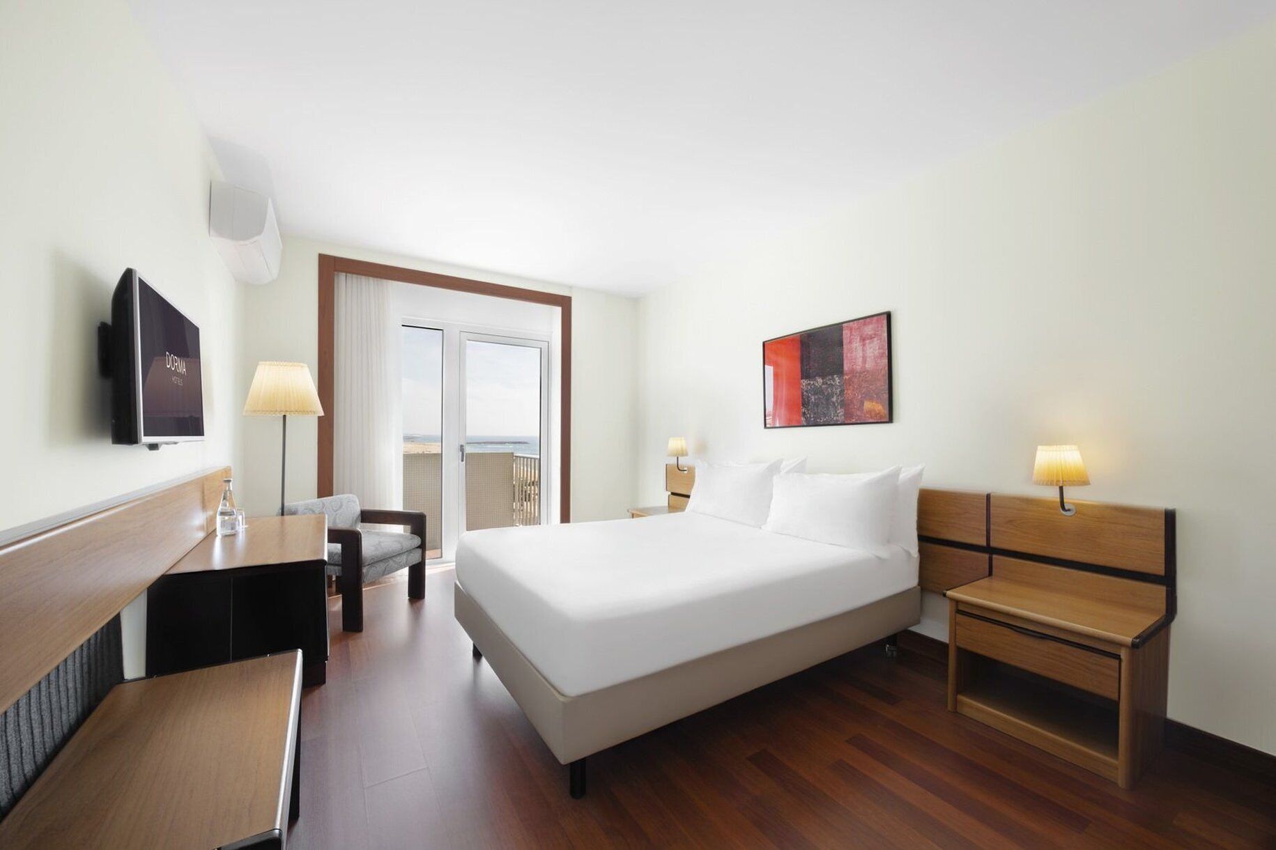 Quarto de hotel com cama, mesas de cabeceira, escrivaninha, cadeira, televisão e vista para o mar através de uma porta de correr.