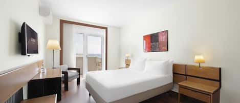 Quarto de hotel com cama, mesas de cabeceira, escrivaninha, cadeira, televisão e vista para o mar através de uma porta de correr.