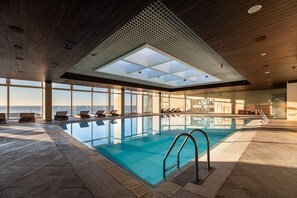 An indoor swimming pool with clear glass walls, a view of the ocean, and a modern ceiling design.