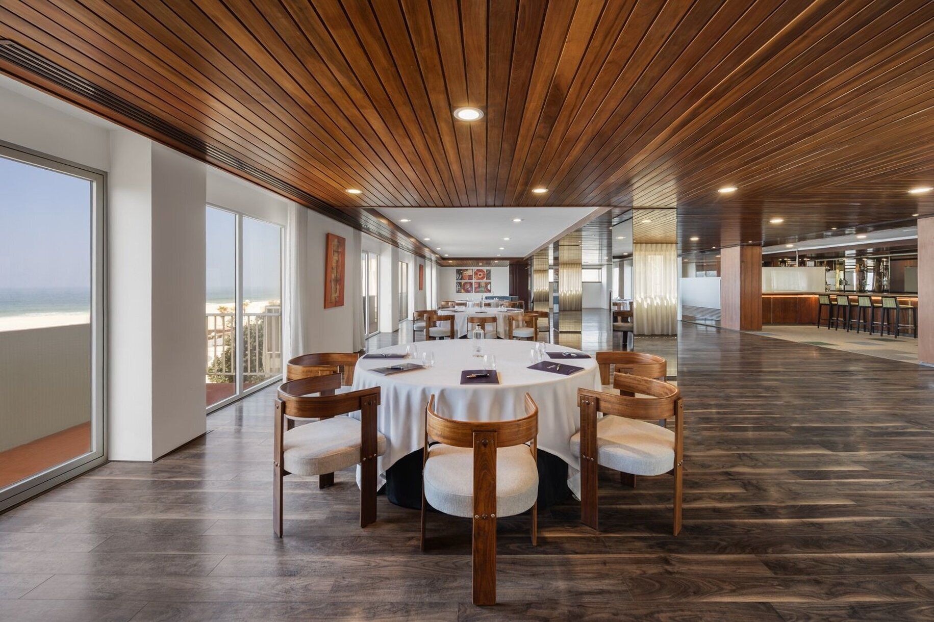 A spacious dining area with a view of the beach, wooden ceiling, and modern furniture.