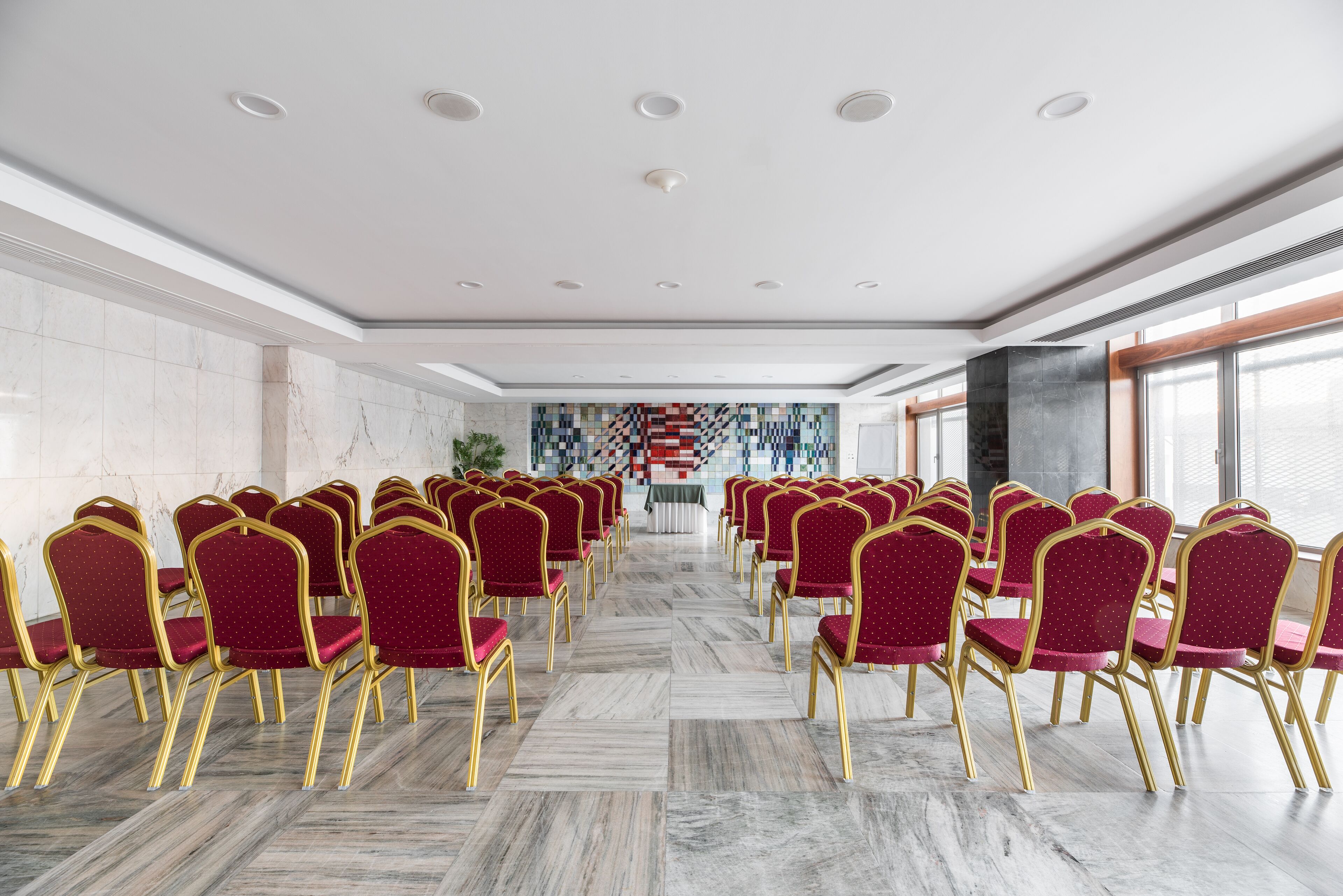 A conference room with rows of red chairs and gold frames, marble flooring, and a large window with a view.