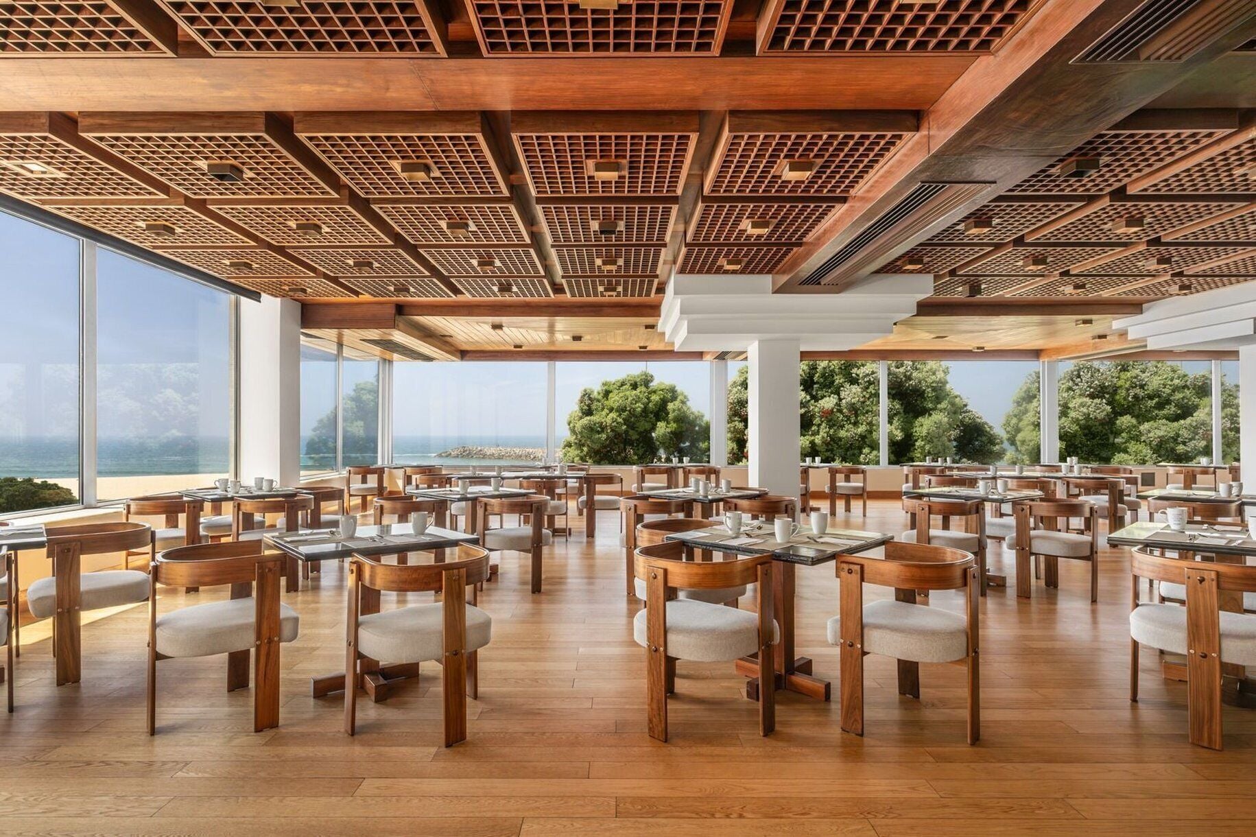 Un vaste espace dédié à la salle à manger, avec des tables et des chaises en bois, de grandes fenêtres offrant une vue sur l’océan et un plafond en bois orné d’un motif à carreaux.