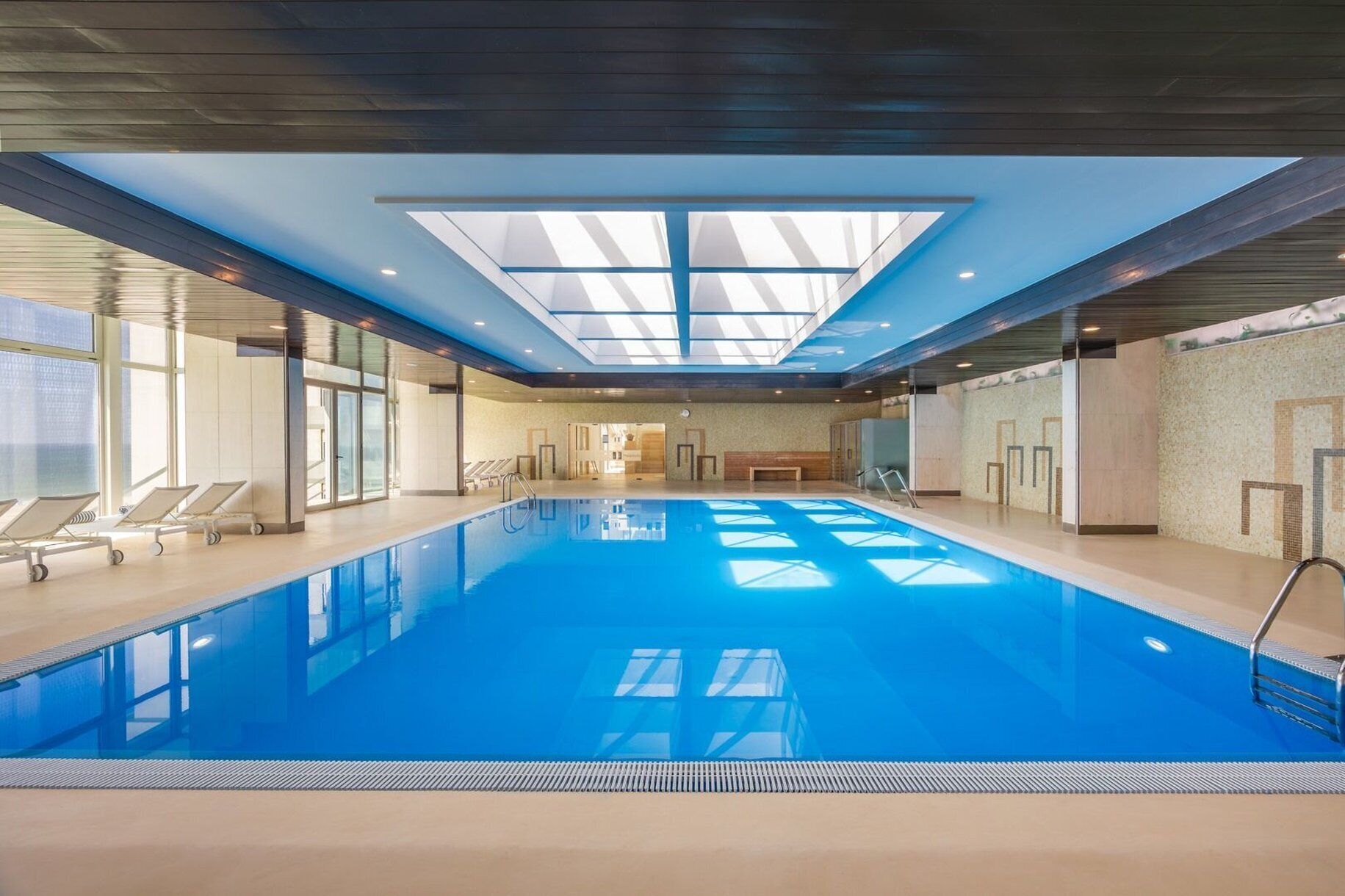A large indoor swimming pool with clear blue water, surrounded by lounge chairs and a skylight ceiling.