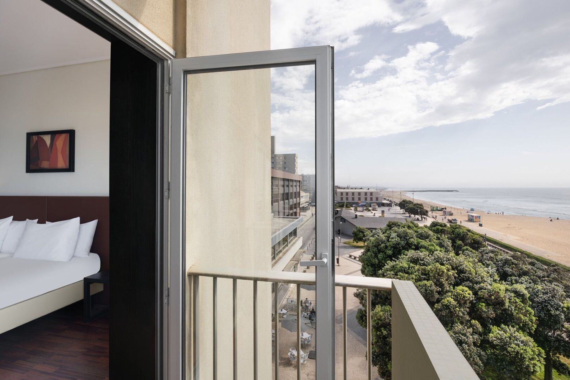 Un balcon avec vue sur la plage et l’océan, un lit avec une literie blanche et un tableau encadré au mur.