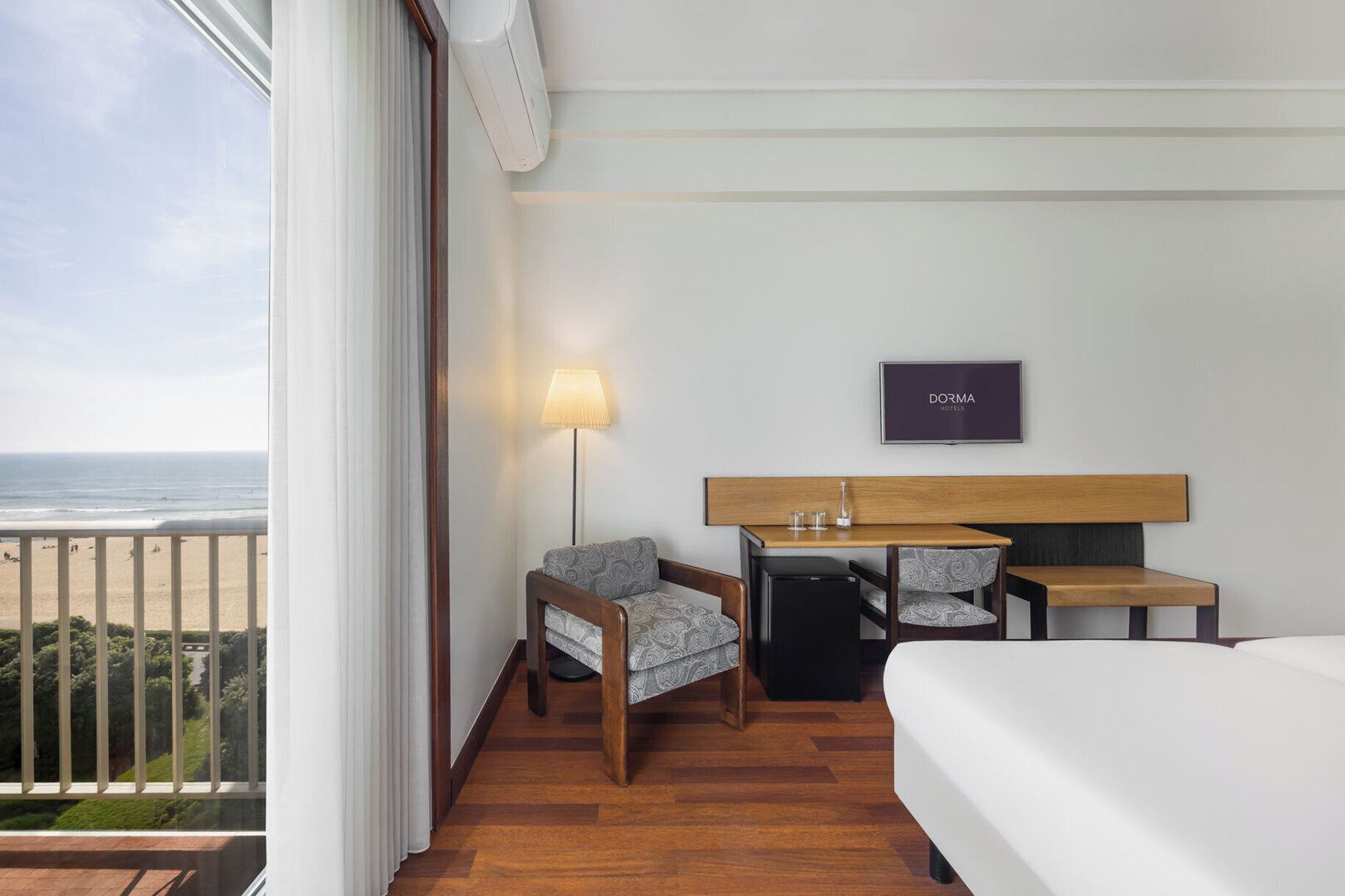 Habitación de hotel con cama, escritorio, silla, lámpara, televisor y vista a la playa.