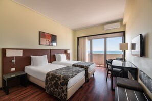 A hotel room with two beds, a desk, a chair, a TV, and a large window with a view of the ocean.