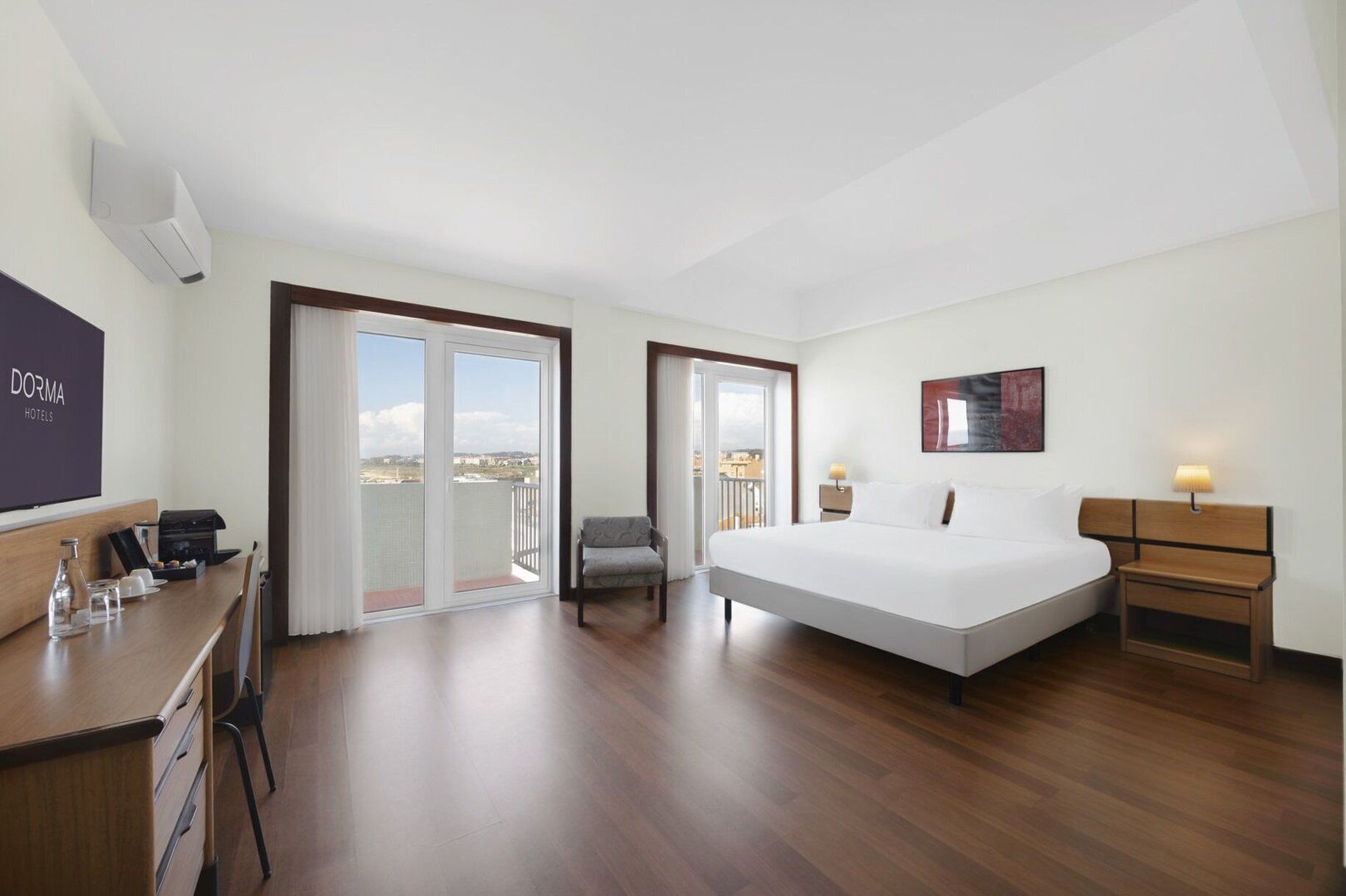 Ein Hotelzimmer mit Bett, Schreibtisch, Stuhl, Nachttisch, Lampe und Blick auf die Stadt durch große Fenster.