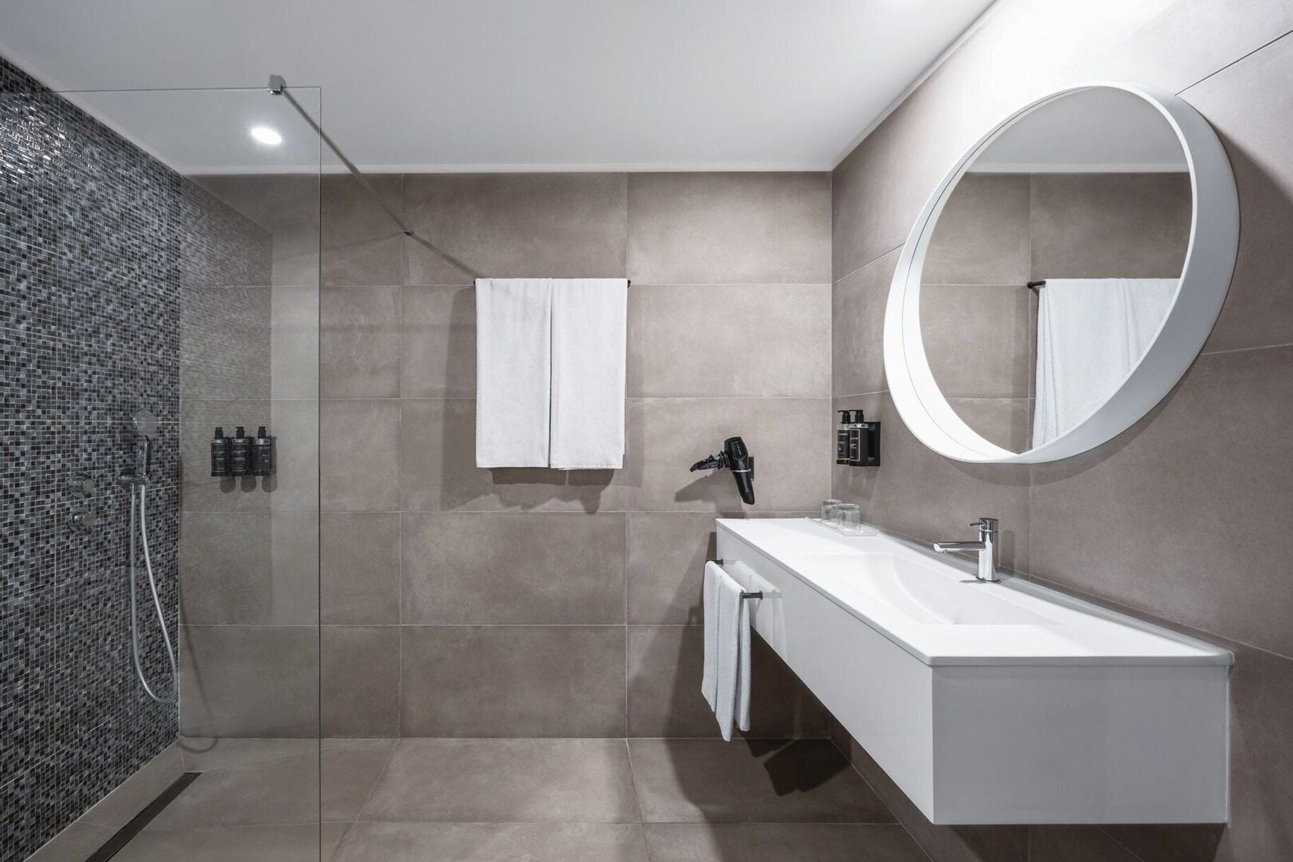 Un baño moderno con ducha de vidrio, tocador blanco, espejo redondo y perchero para toallas.