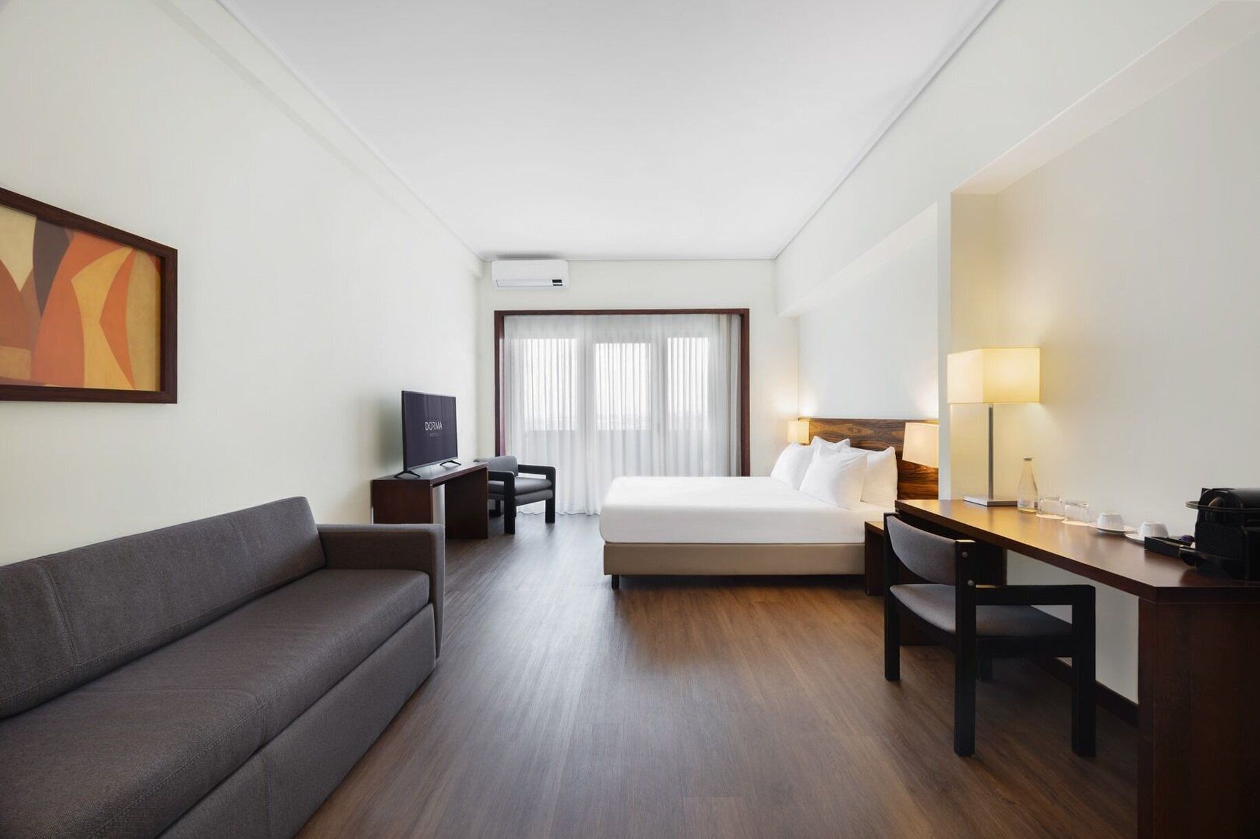 A hotel room with a bed, a desk, a chair, a sofa, a TV, and a lamp.